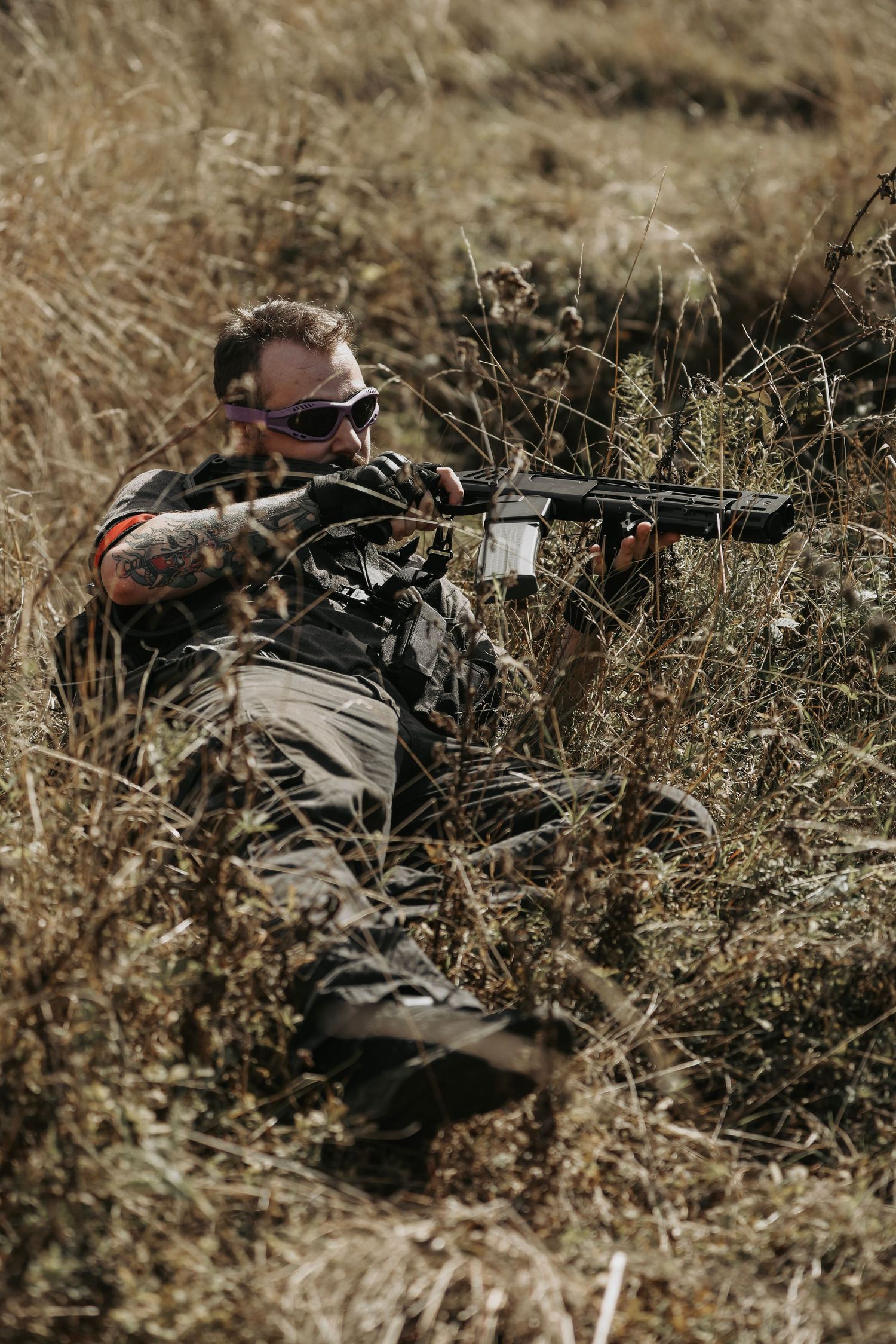 Tattooed soldier in tactical gear aiming rifle while concealed in dry grass field during military training exercise.