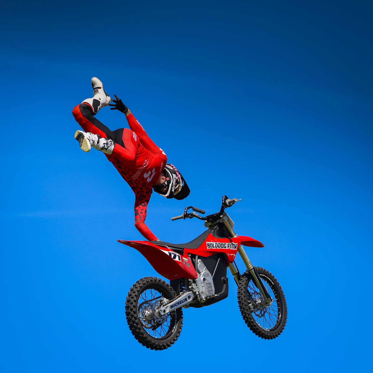 Freestyle motocross rider in red gear performing a backflip trick on a red dirt bike against blue sky. Photo Taken by AJCoatesPhotography