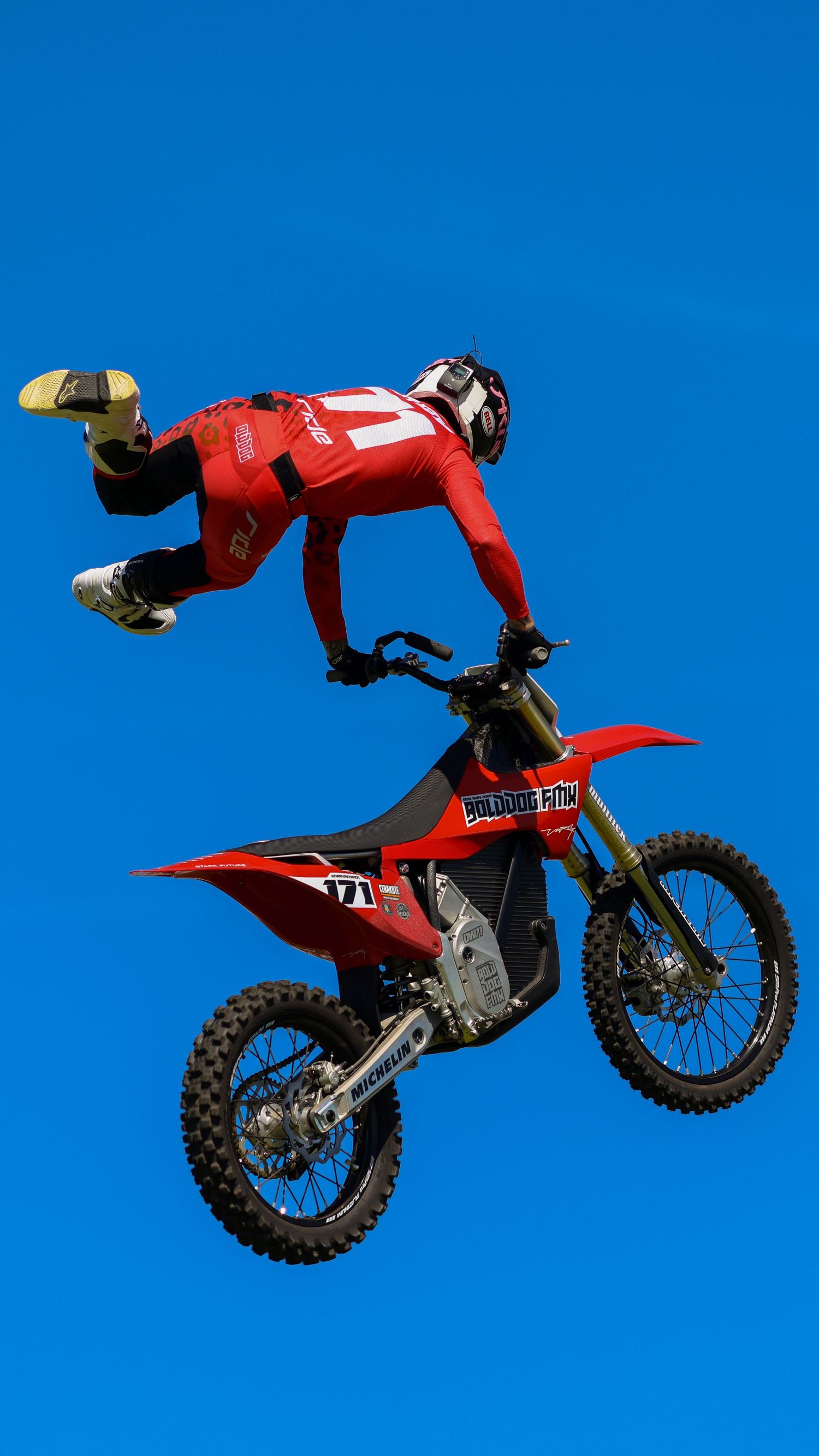 FMX rider in red jersey number 171 executing a Superman trick on a red dirt bike against blue sky. Photo Taken by AJCoatesPhotography