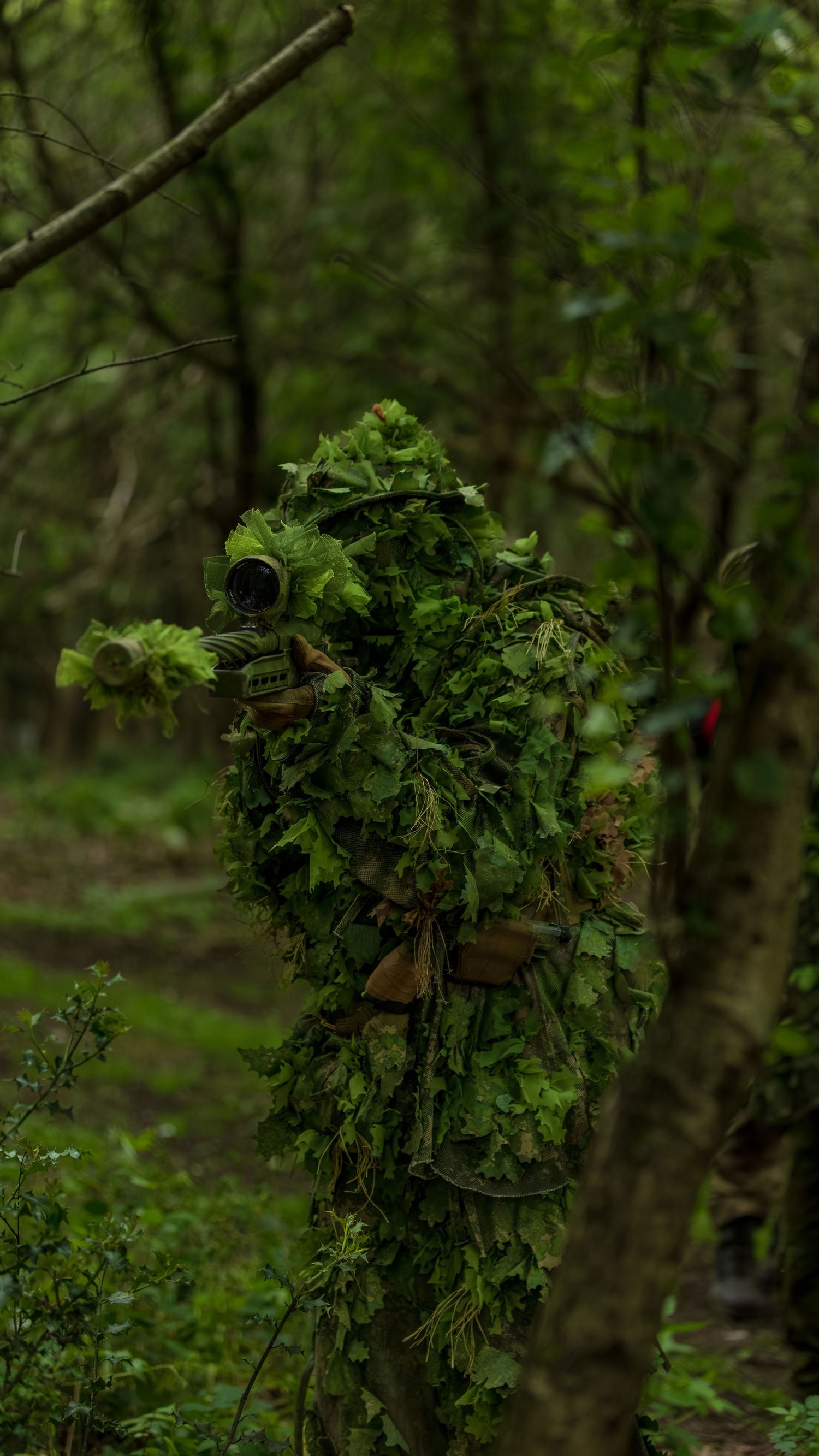 Person in full ghillie suit camouflaged among green foliage and trees in a forest. Photo Taken by AJCoatesPhotography