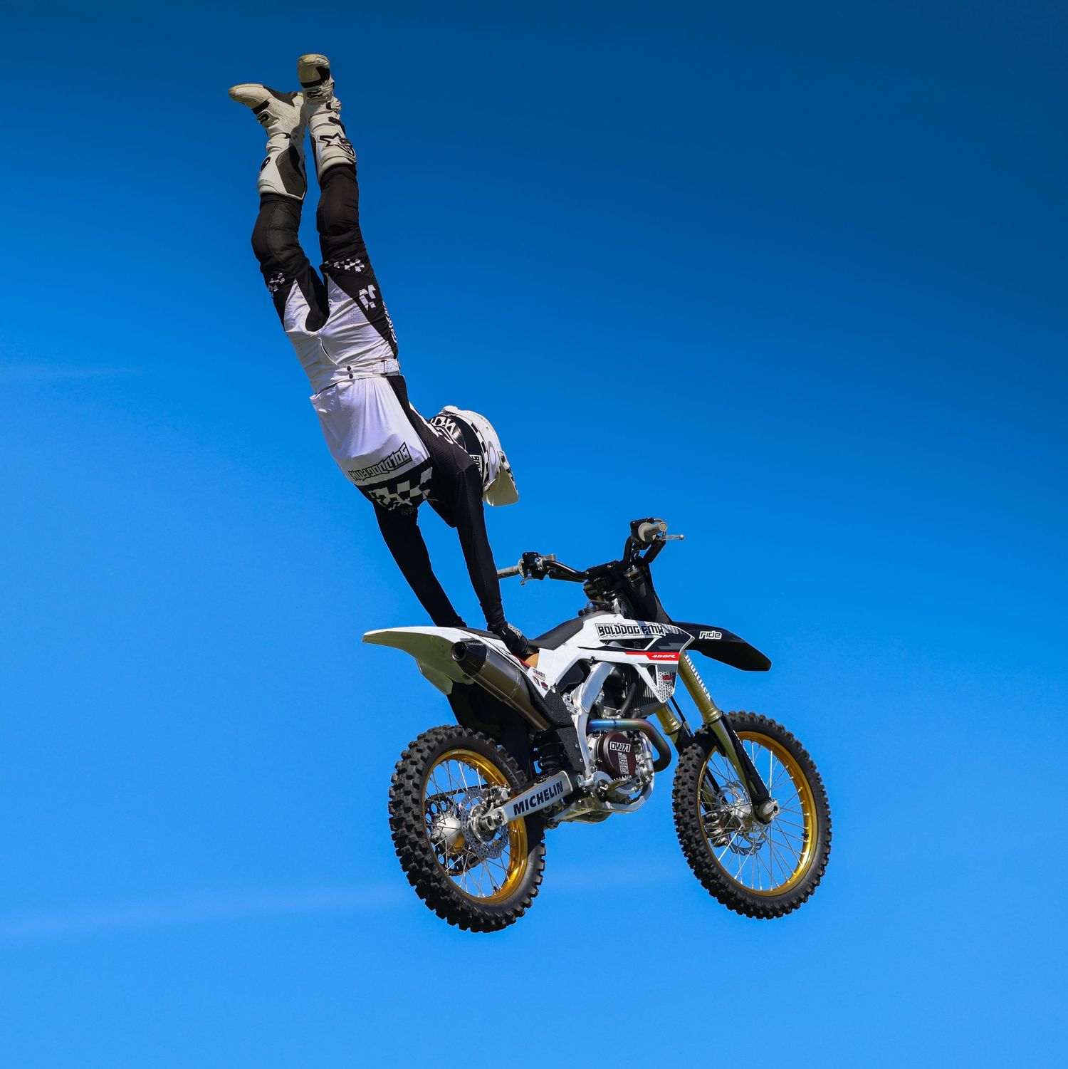 Freestyle motocross rider performing a headstand trick on a dirt bike mid-air against a clear blue sky. Photo Taken by AJCoatesPhotography