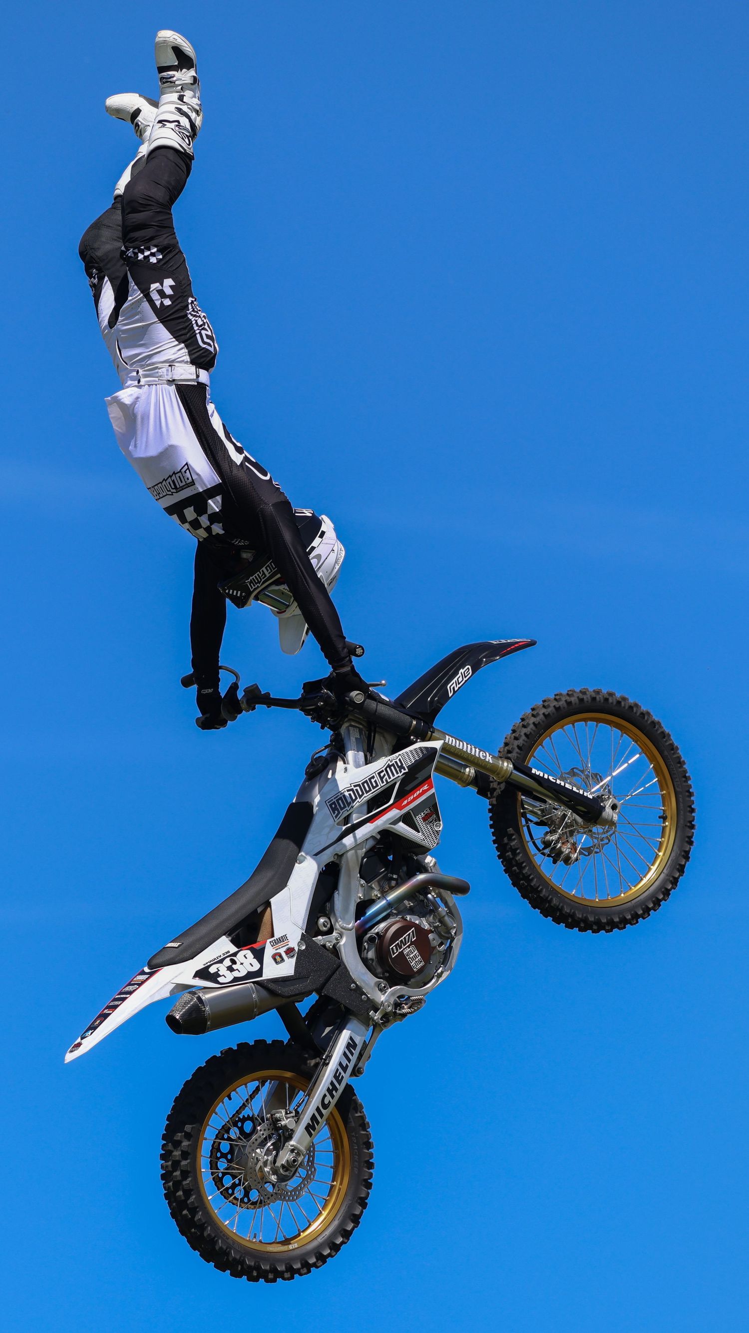 FMX rider performing a vertical inversion trick on a white and gold dirt bike against a bright blue sky. Photo Taken by AJCoatesPhotography