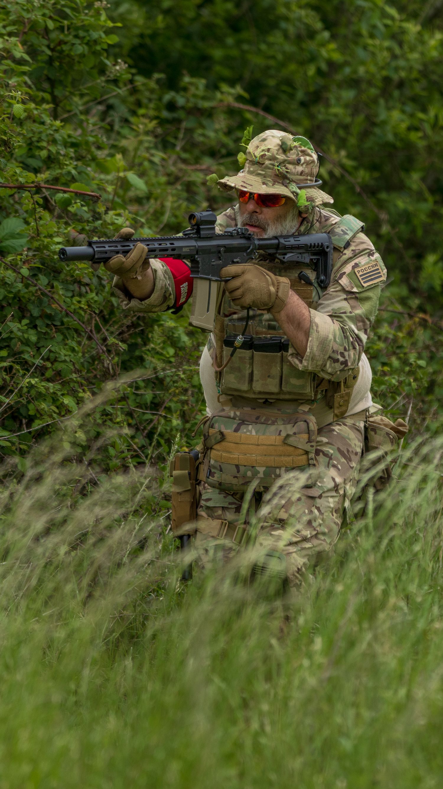 Armed soldier in multicam tactical gear aiming rifle through dense green vegetation during outdoor military exercise. Photo Taken by AJCoate