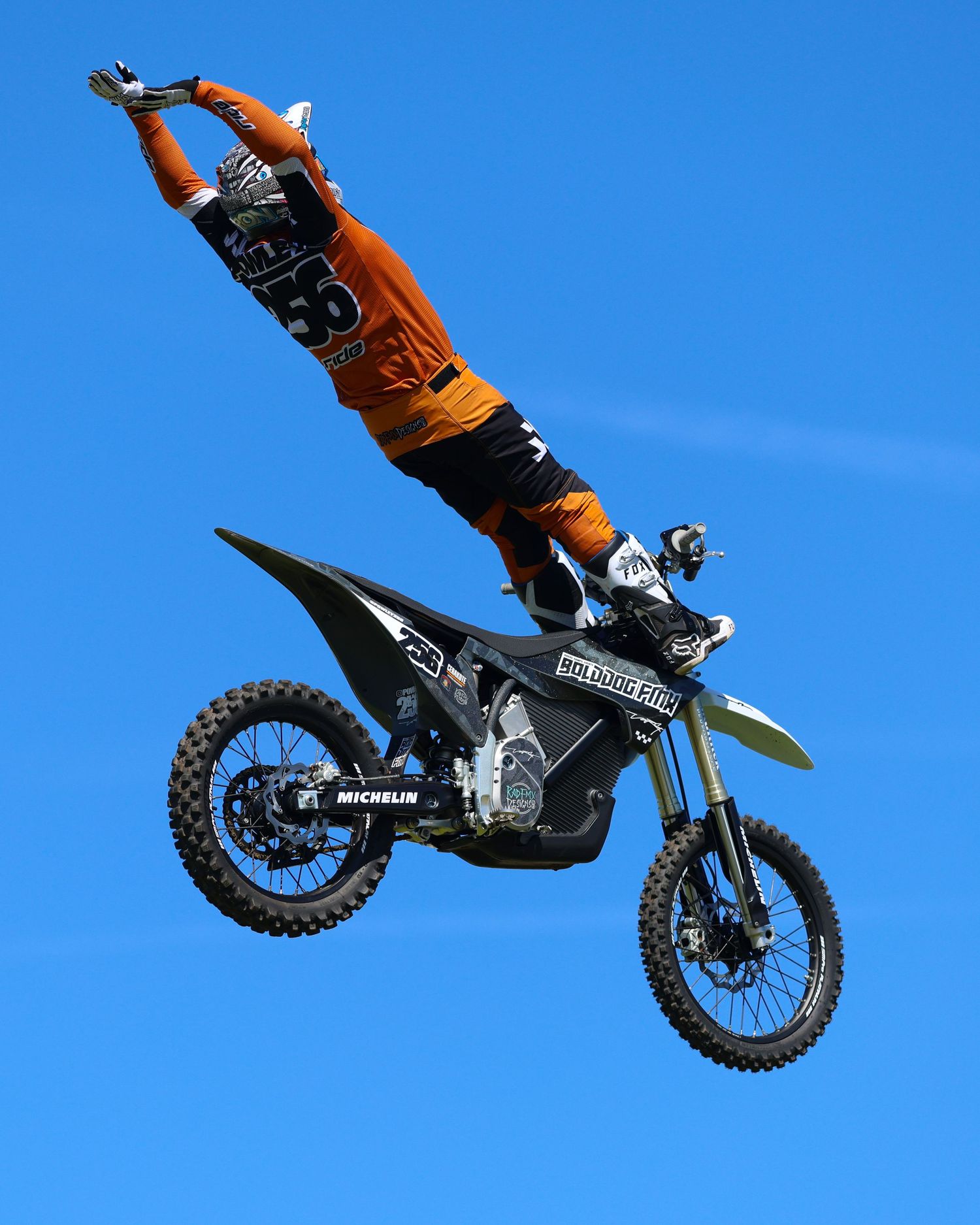 Freestyle motocross rider in orange gear standing upright on a dark dirt bike during a high aerial jump. Photo Taken by AJCoatesPhotography