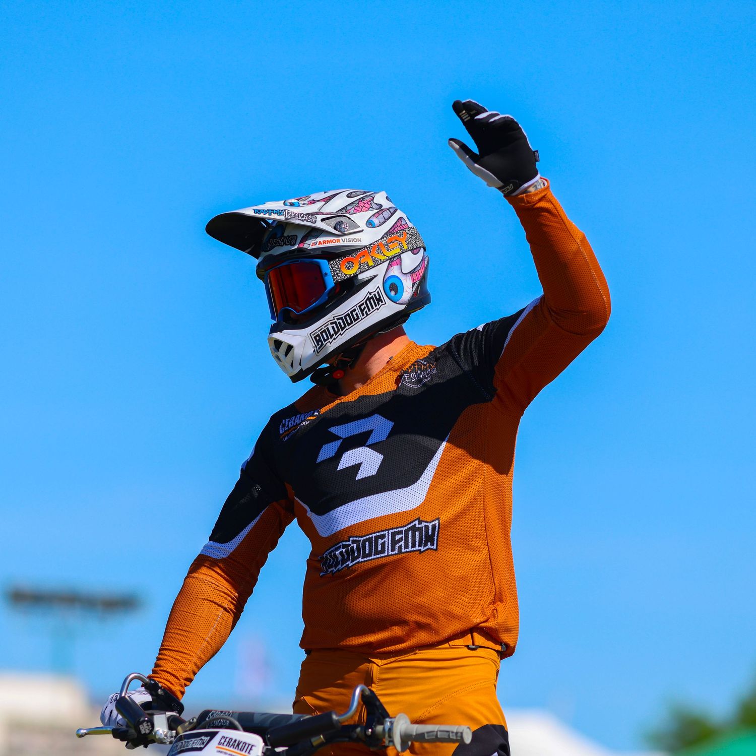 Motocross rider in orange jersey waving hand while on bike under clear blue sky. Photo Taken by AJCoatesPhotography