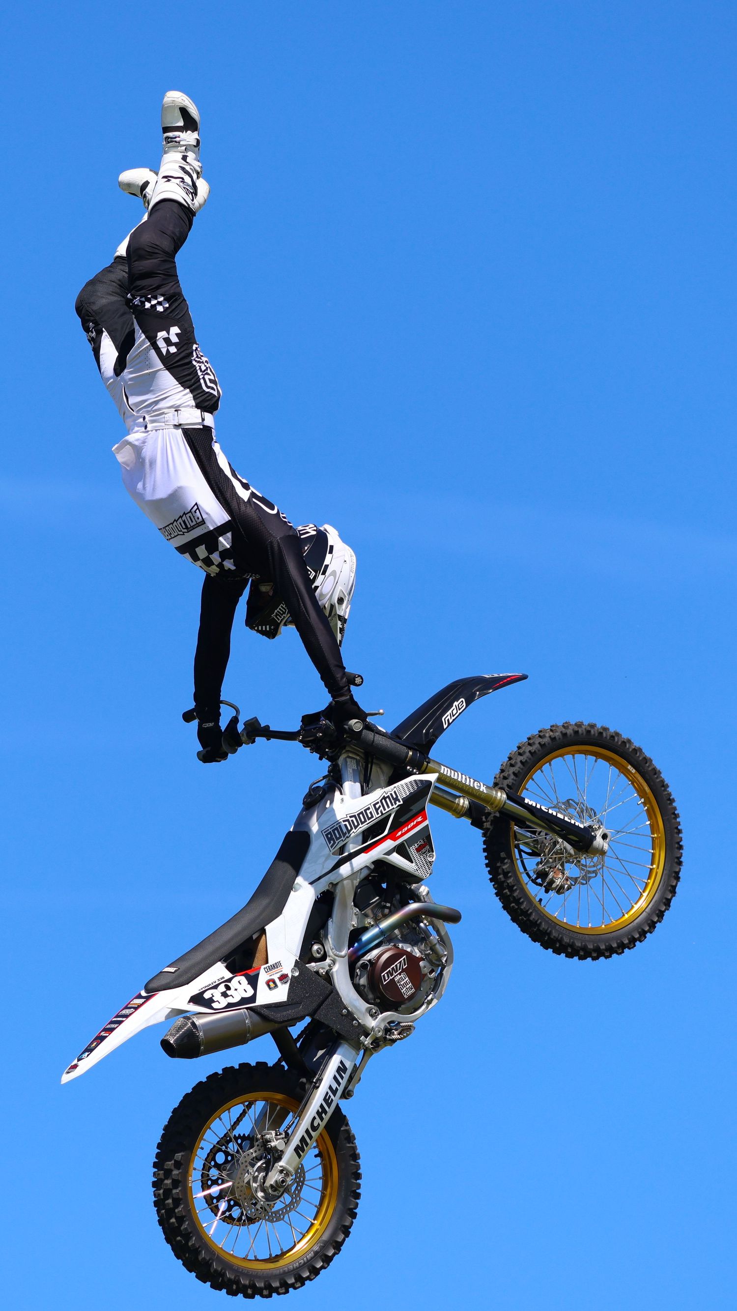 Freestyle motocross rider executing a vertical inversion stunt on a white and gold dirt bike in blue sky. Photo Taken by AJCoatesPhotography
