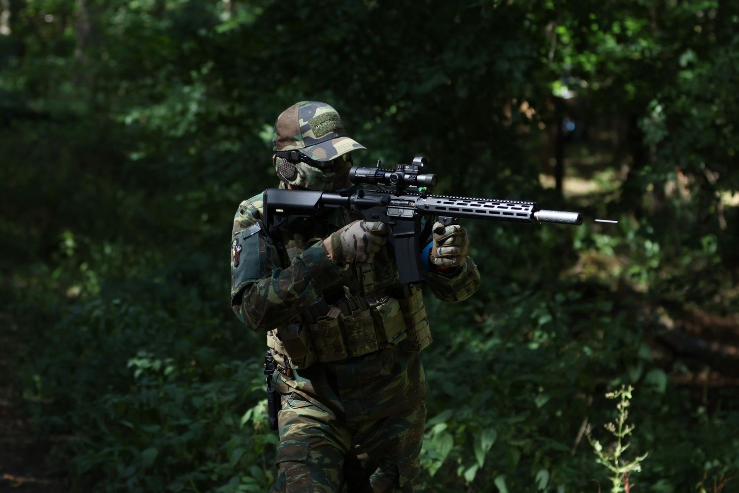 Soldier in camouflage uniform aiming assault rifle with scope in dense forest during military training exercise.