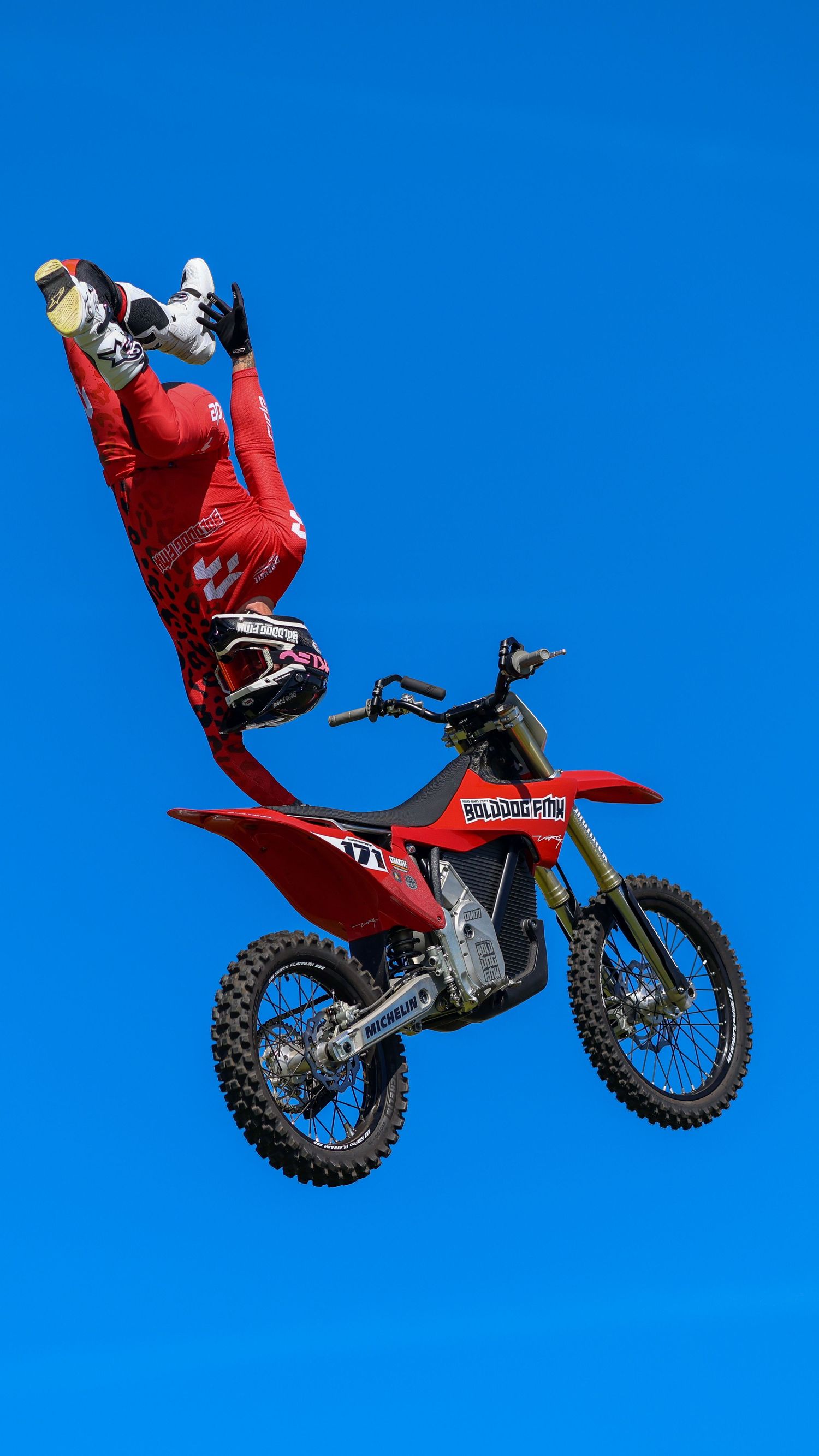 Freestyle motocross rider in red gear performing an inverted trick high in the air against a clear blue sky. Photo Taken by AJCoatesPhotogra