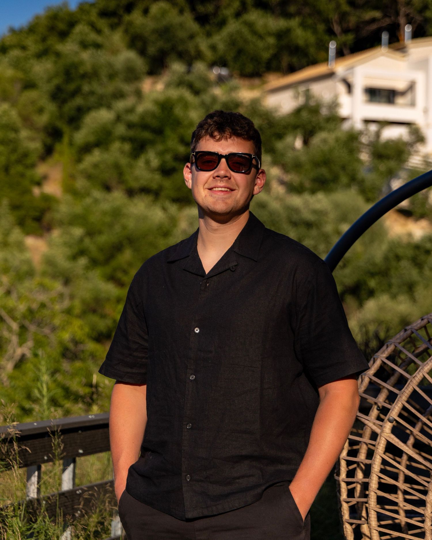 Young man in black shirt and sunglasses smiling outdoors near lush greenery. Photo Taken by AJCoatesPhotography