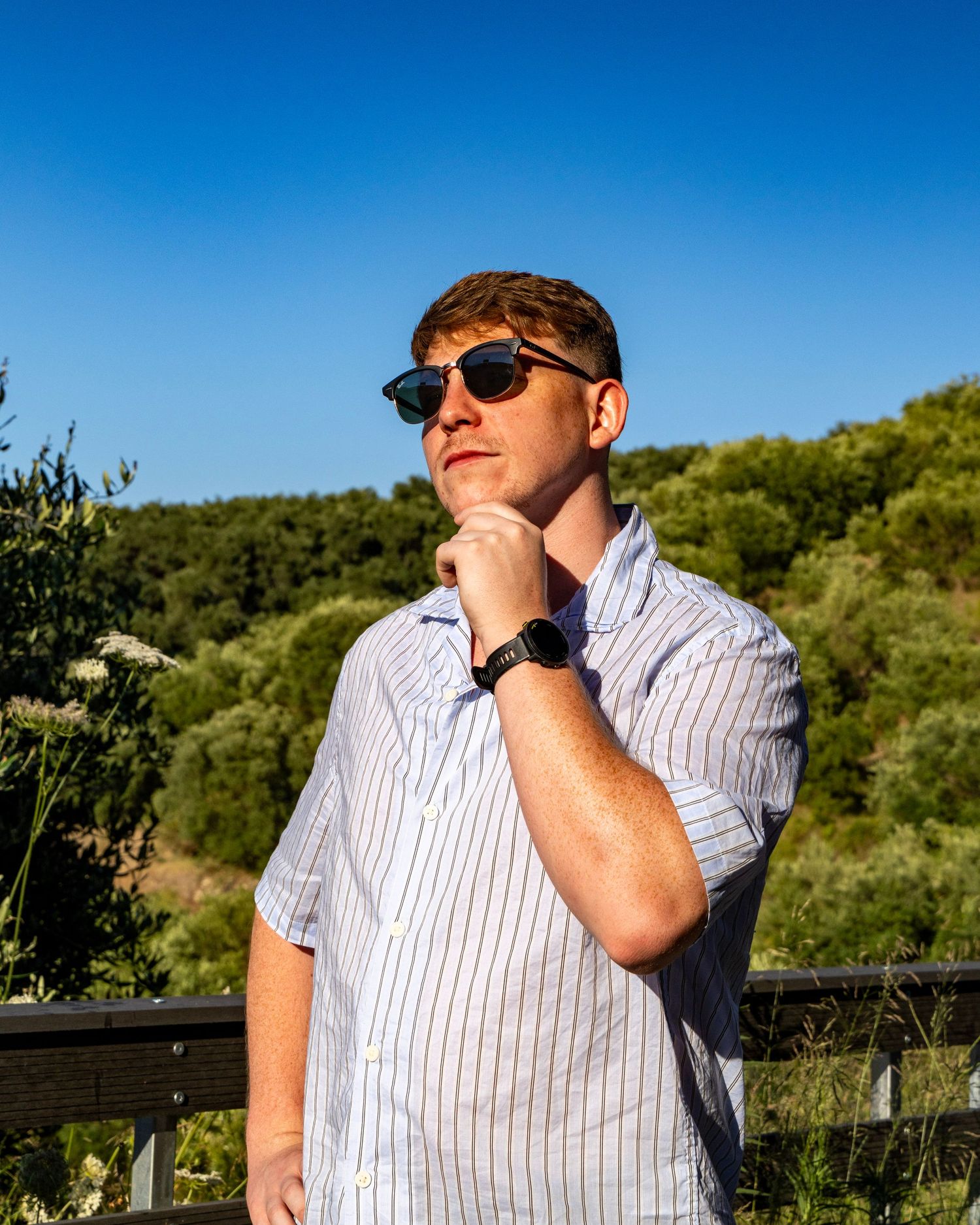 Young man in striped shirt and sunglasses posing outdoors against green hillside. Photo Taken by AJCoatesPhotography