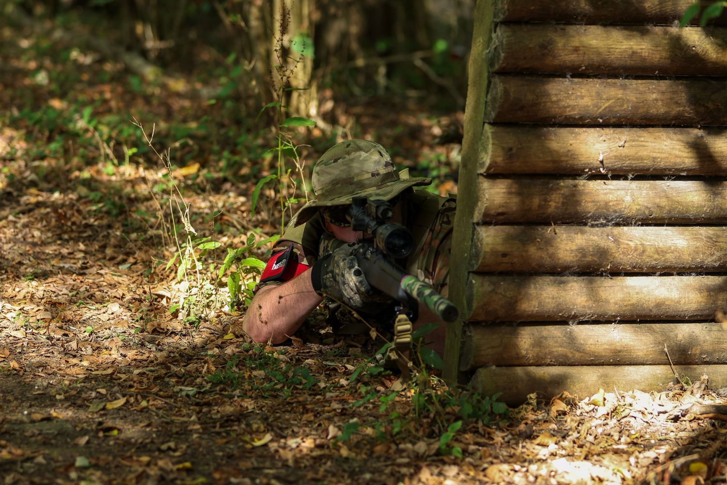 A paintball player crouches behind a wooden log bunker wearing camouflage gear and protective mask in a forest arena.