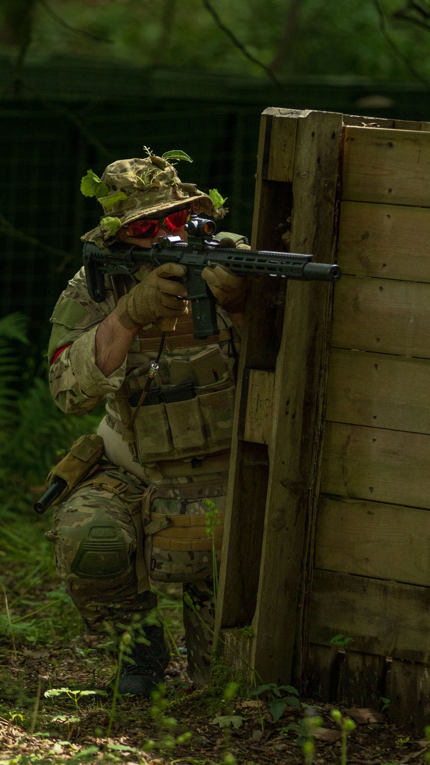 Tactical soldier in multicam gear crouching behind wooden barrier aiming rifle with red-lens goggles in forest. Photo Taken by AJCoatesPhoto