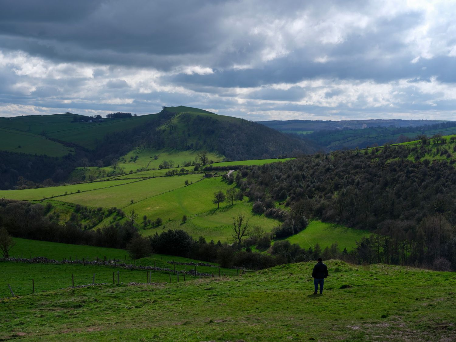 Lone hiker overlooking lush green rolling hills valley under dramatic cloudy sky. Photo Taken by AJCoatesPhotography