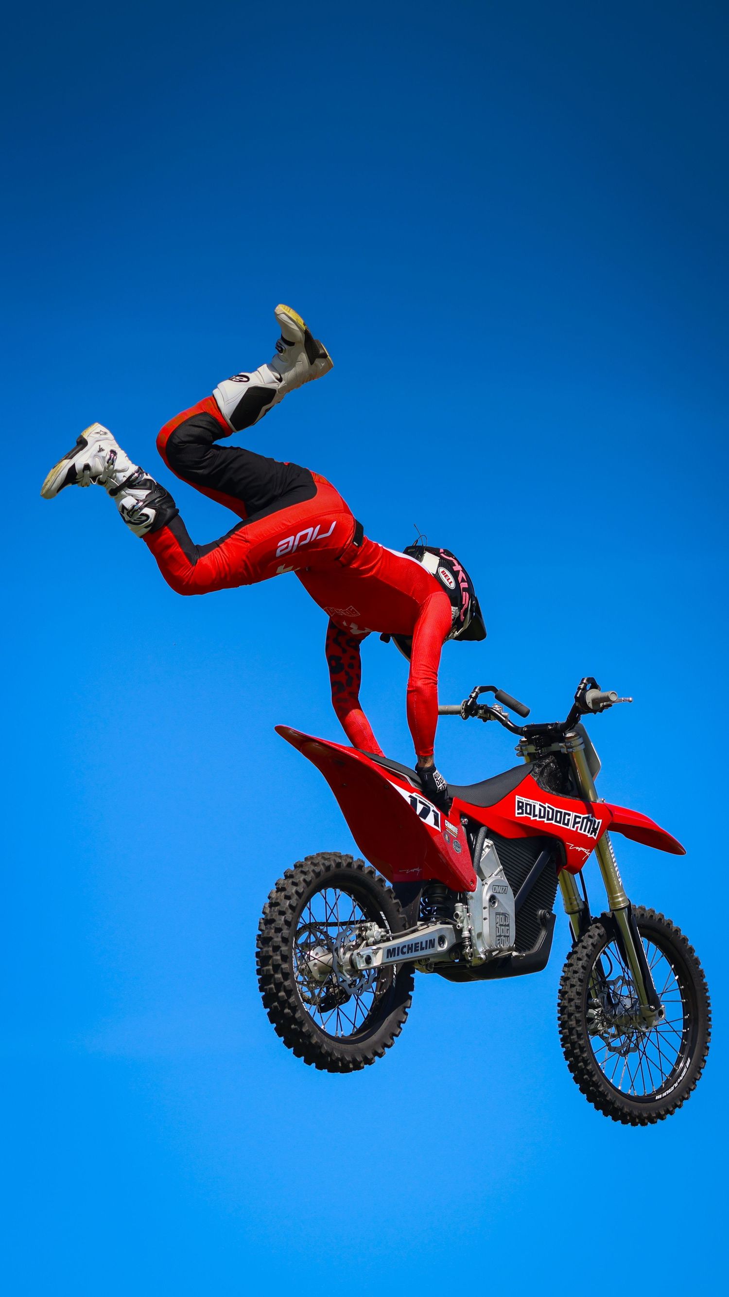 Freestyle motocross rider in red standing on seat of airborne red dirt bike against blue sky. Photo Taken by AJCoatesPhotography