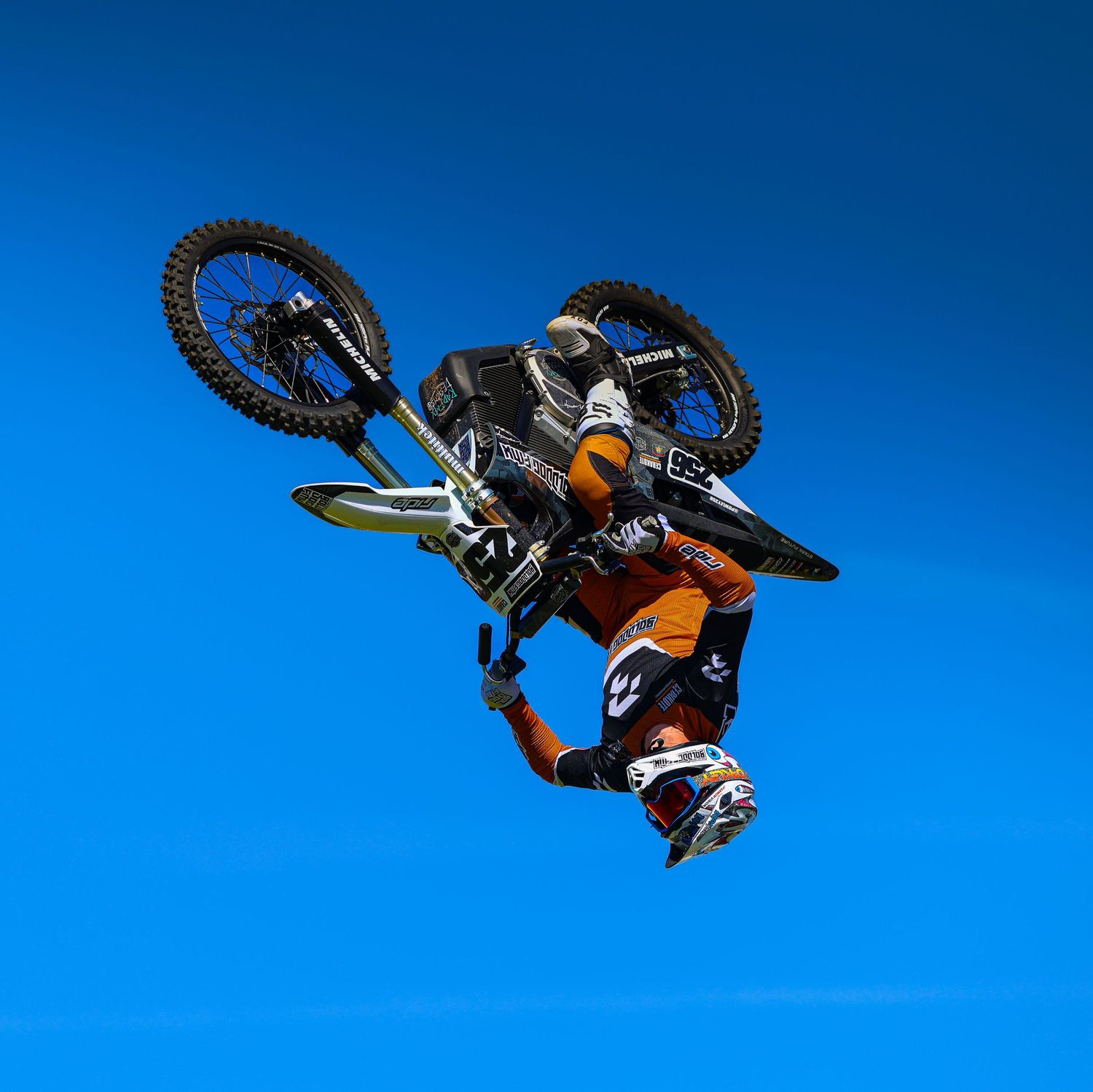 Motocross rider in orange gear performing a dramatic upside-down aerial flip trick against a blue sky. Photo Taken by AJCoatesPhotography