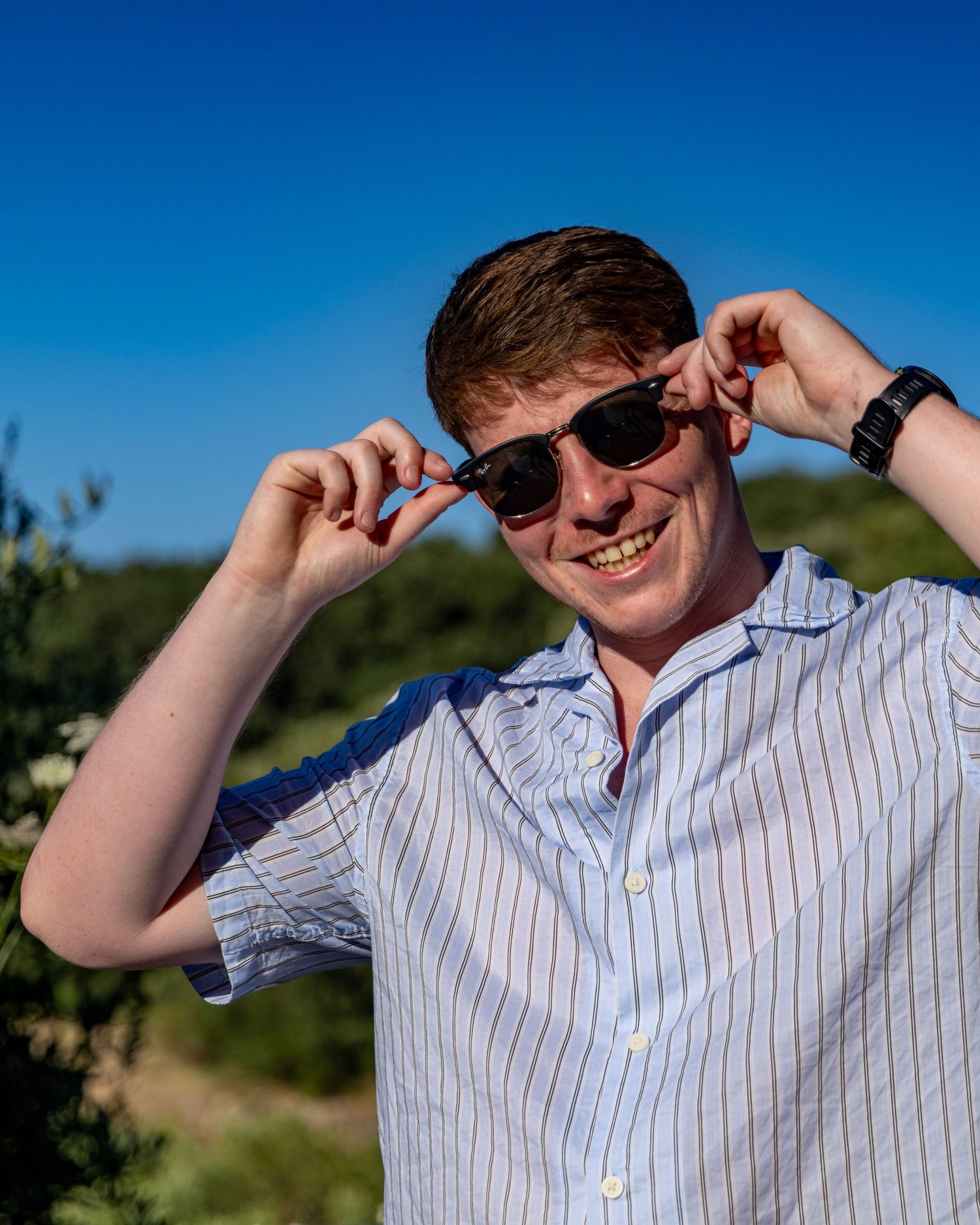 Young man smiling outdoors adjusting black sunglasses, wearing striped shirt under clear blue sky. Photo Taken by AJCoatesPhotography
