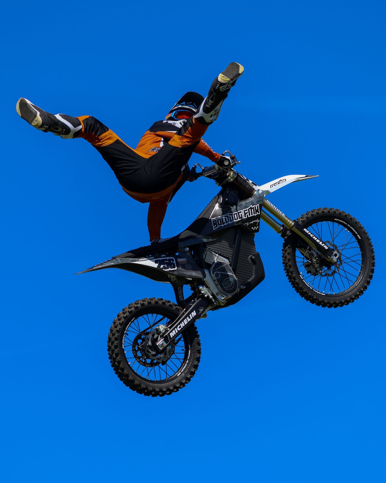 Motocross athlete in orange gear performing a daring aerial stunt on a black dirt bike against blue sky. Photo Taken by AJCoatesPhotography