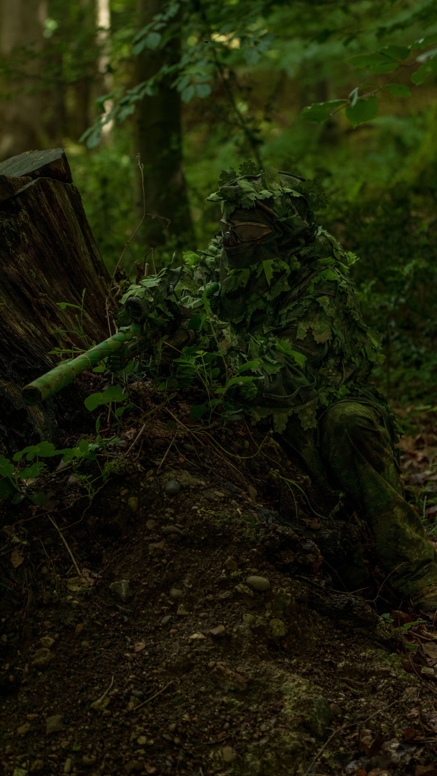 Sniper in detailed ghillie suit blending into forest floor near mossy log in woodland. Photo Taken by AJCoatesPhotography
