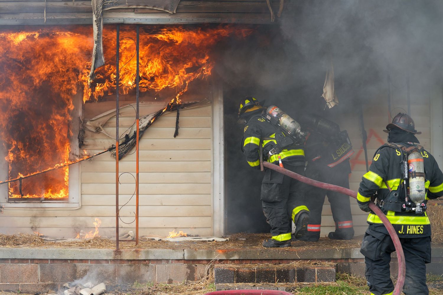 Firefighters advance a hose into a smoke-filled house as flames burn through a nearby window.