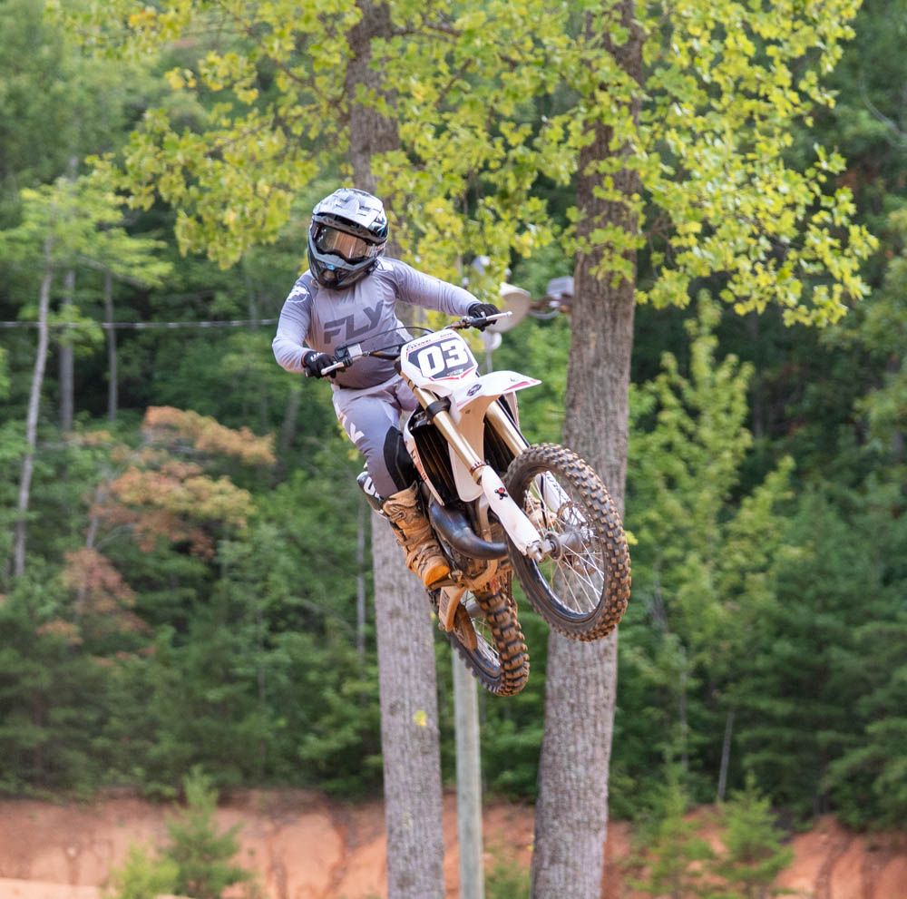 Motocross rider launching high over a jump with trees in the background.