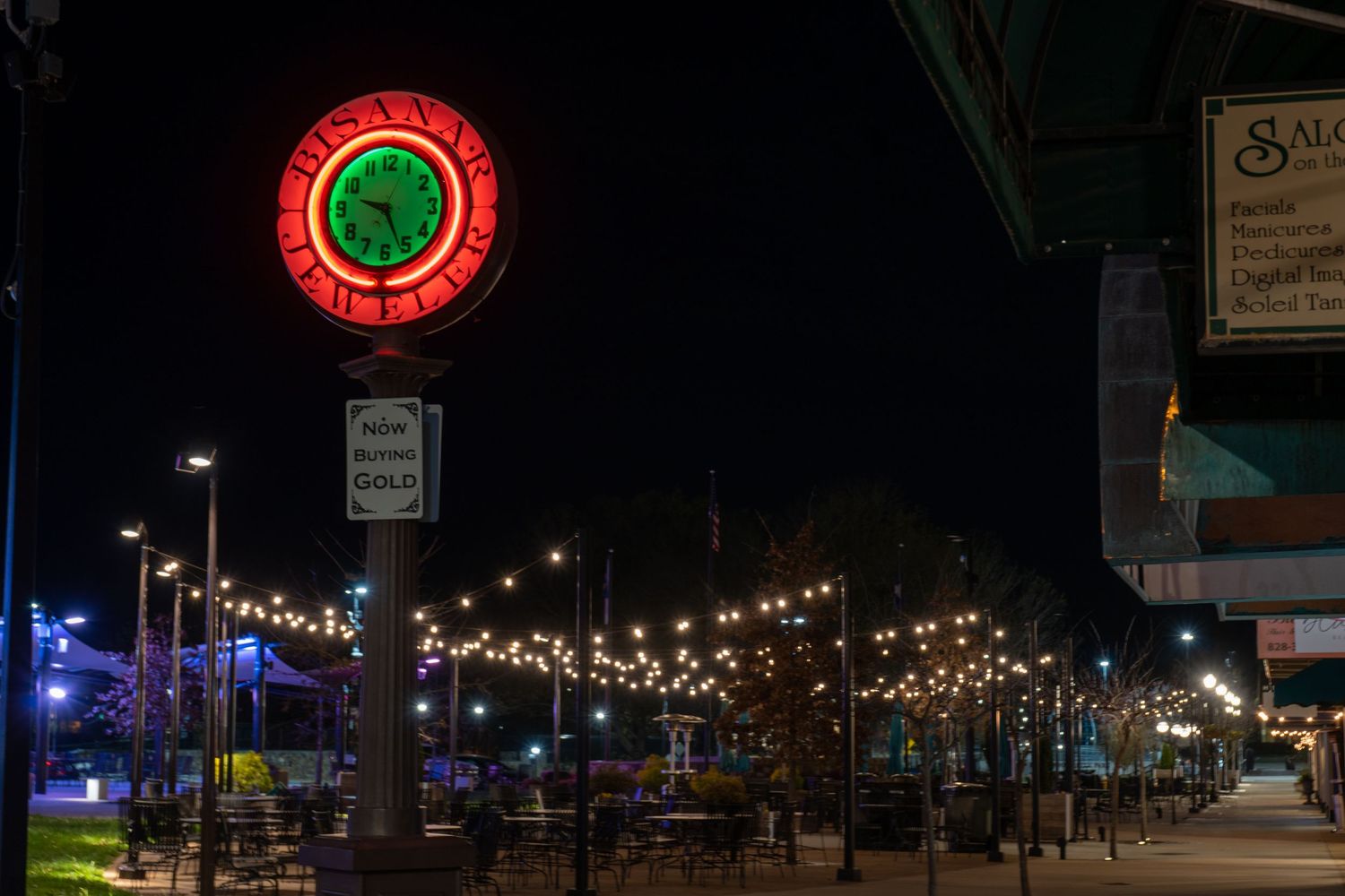 Neon clock overlooking a downtown street illuminated by string lights at night.