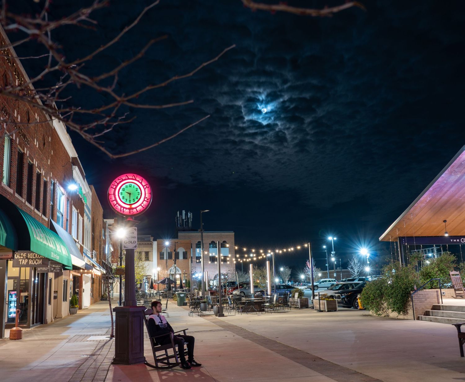 Downtown street at night beneath string lights with the moon glowing through dramatic clouds.