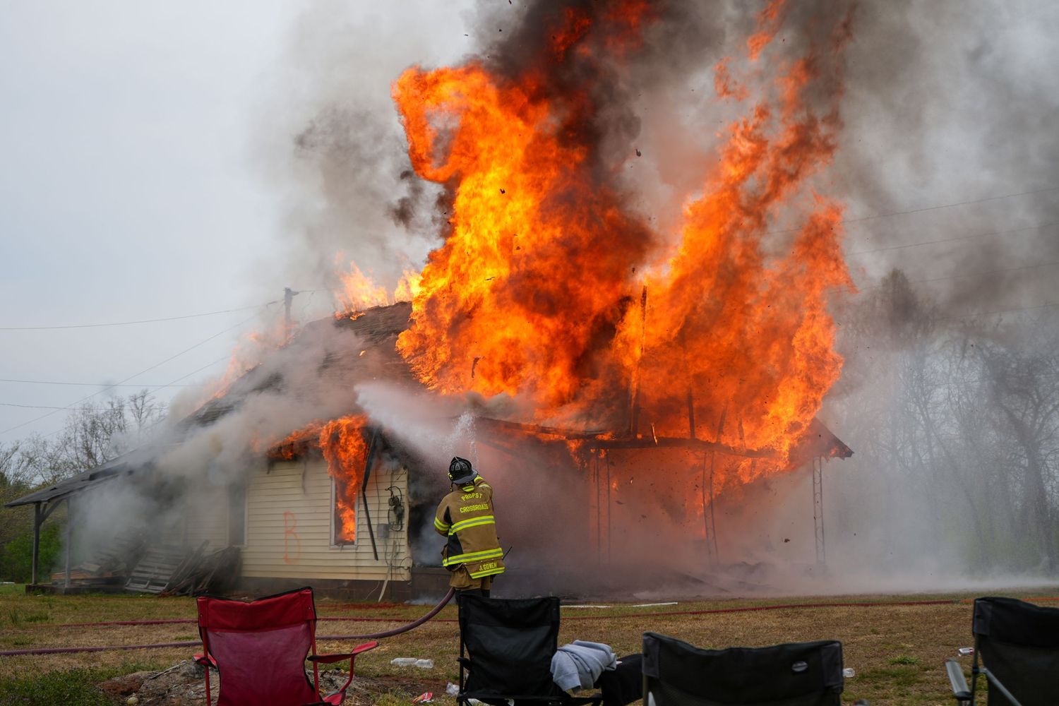 Firefighter sprays water toward a house engulfed in towering flames and heavy smoke.