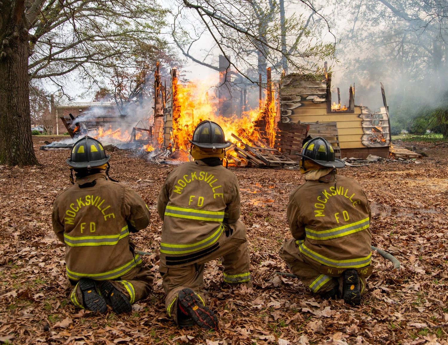 Three firefighters kneel and watch a burning structure during a fire training exercise.