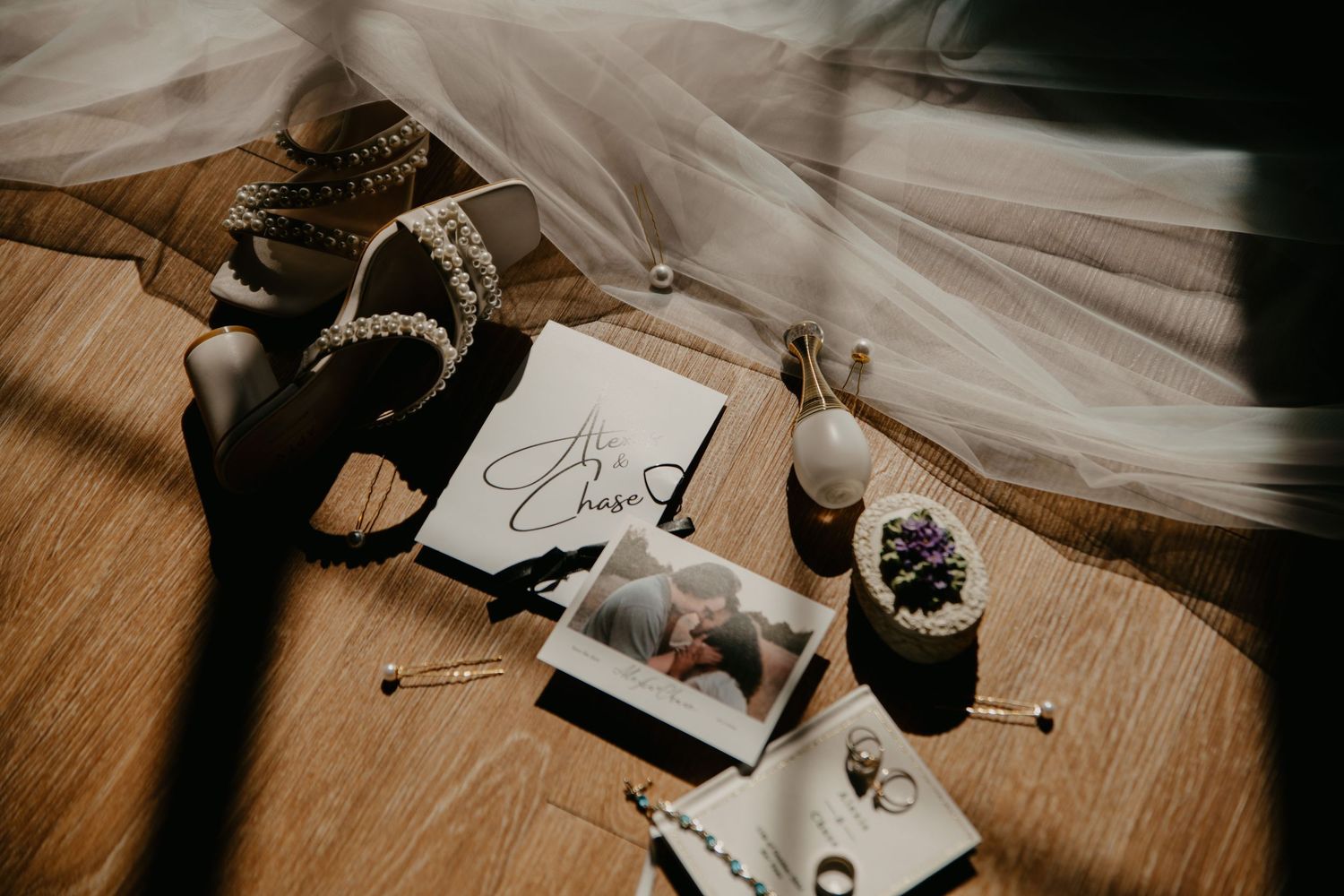 Wedding flat lay with veil, heels, guest book, rings, and bridal accessories on wooden floor.