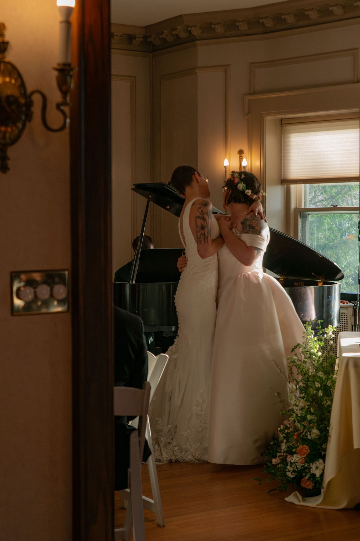 Two brides sharing an emotional embrace near a grand piano in an elegantly decorated indoor venue.
