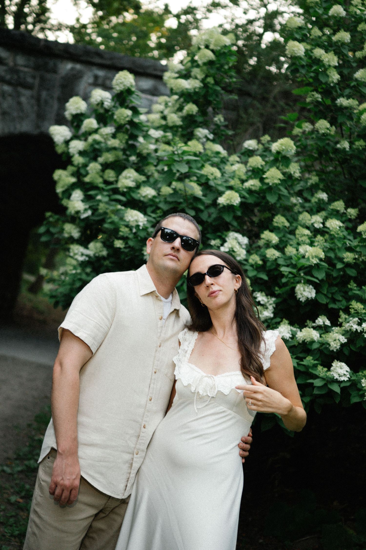 A couple in sunglasses poses before white hydrangea bushes, wearing neutral tones, in a romantic outdoor setting.