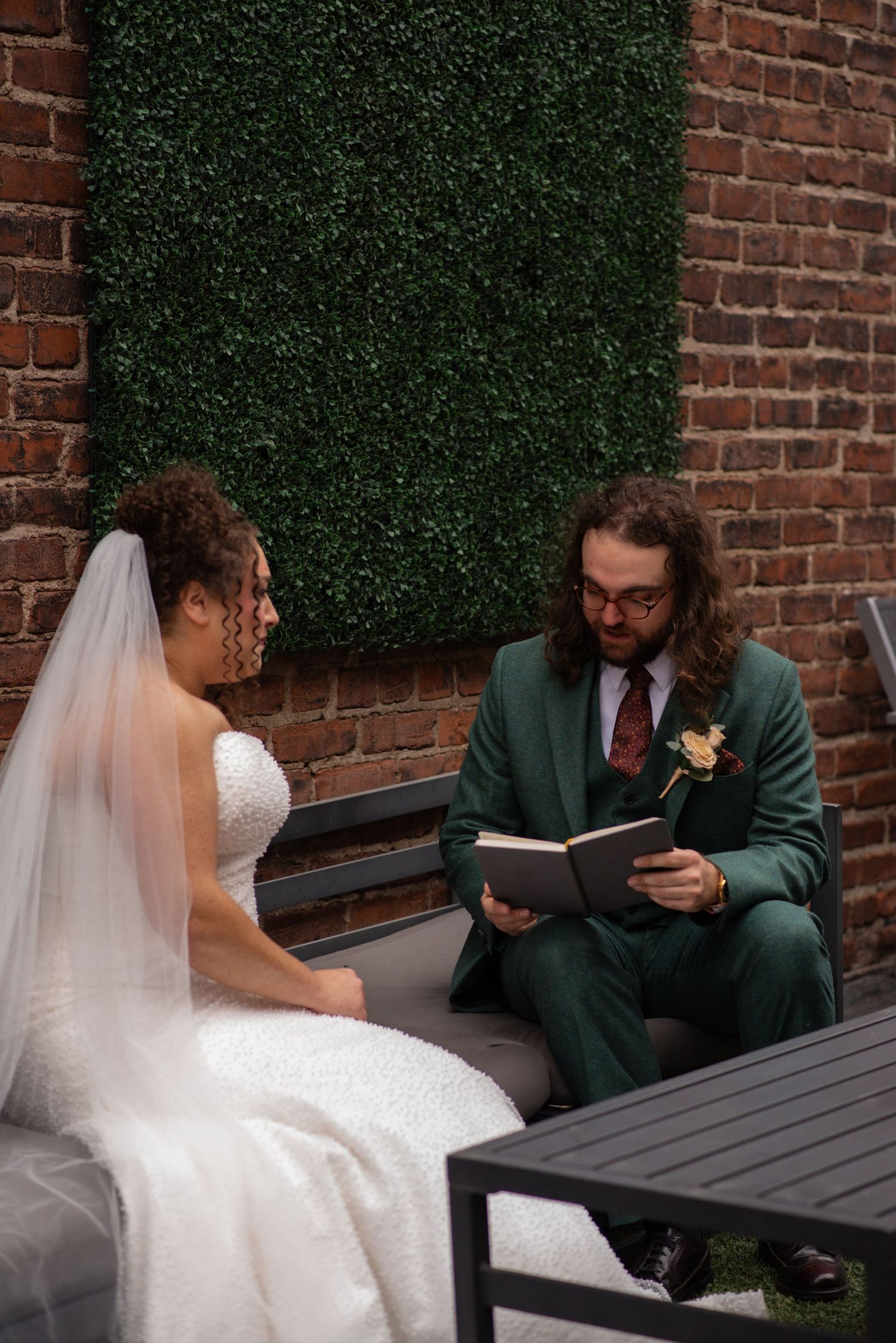 Groom in green suit reads vows from book to bride in white dress, seated outdoors against brick wall with greenery backdrop.