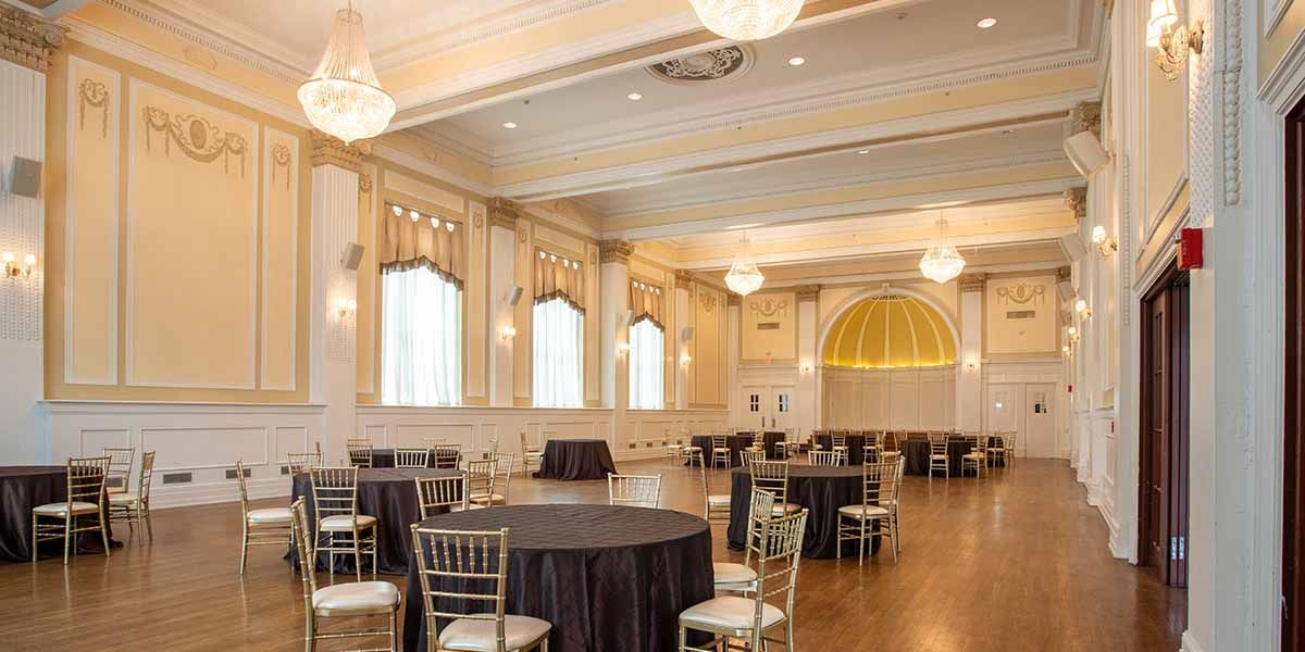 Elegant ballroom with ornate white walls, crystal chandeliers and round tables set up for a formal event.