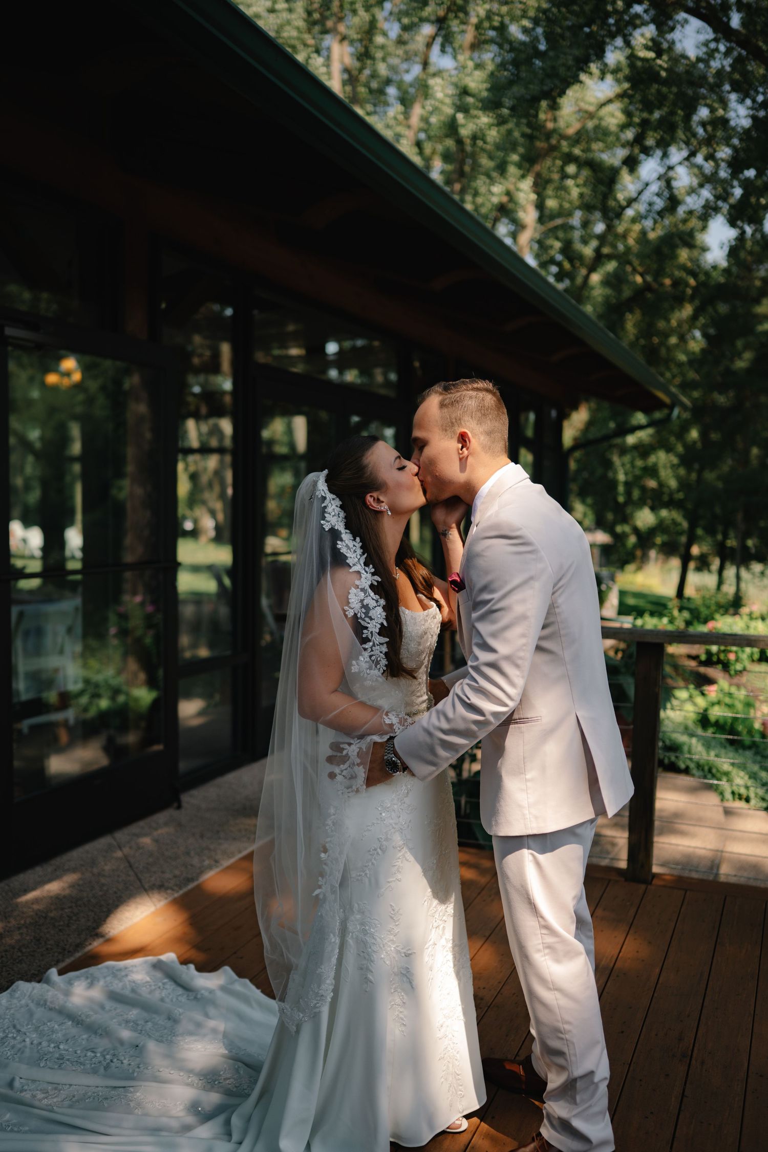 A bride and groom share a romantic kiss on a wooden deck surrounded by lush green trees on their wedding day.