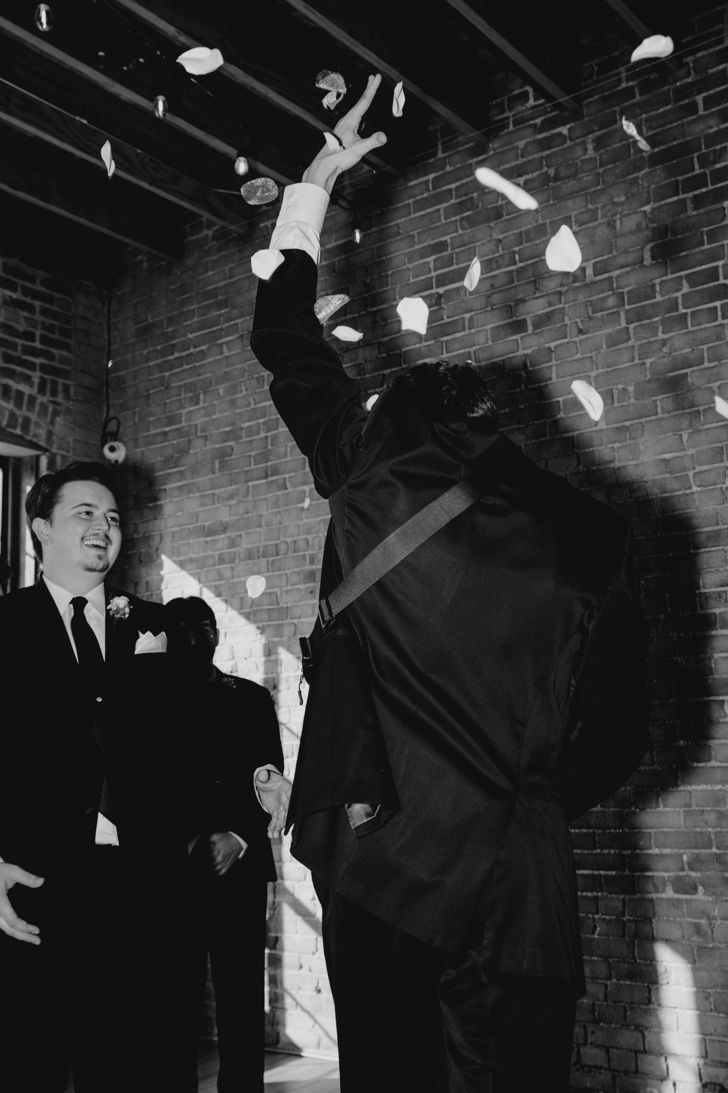 Black and white photo of groom tossing flower petals at a wedding celebration indoors.