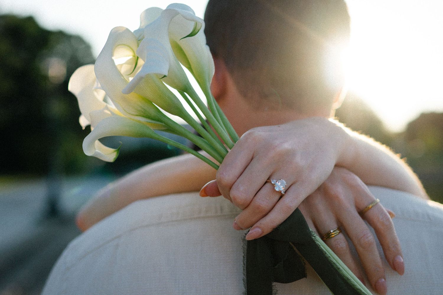Bride holds white calla lily bouquet with diamond engagement ring visible while embracing groom in golden sunlight.