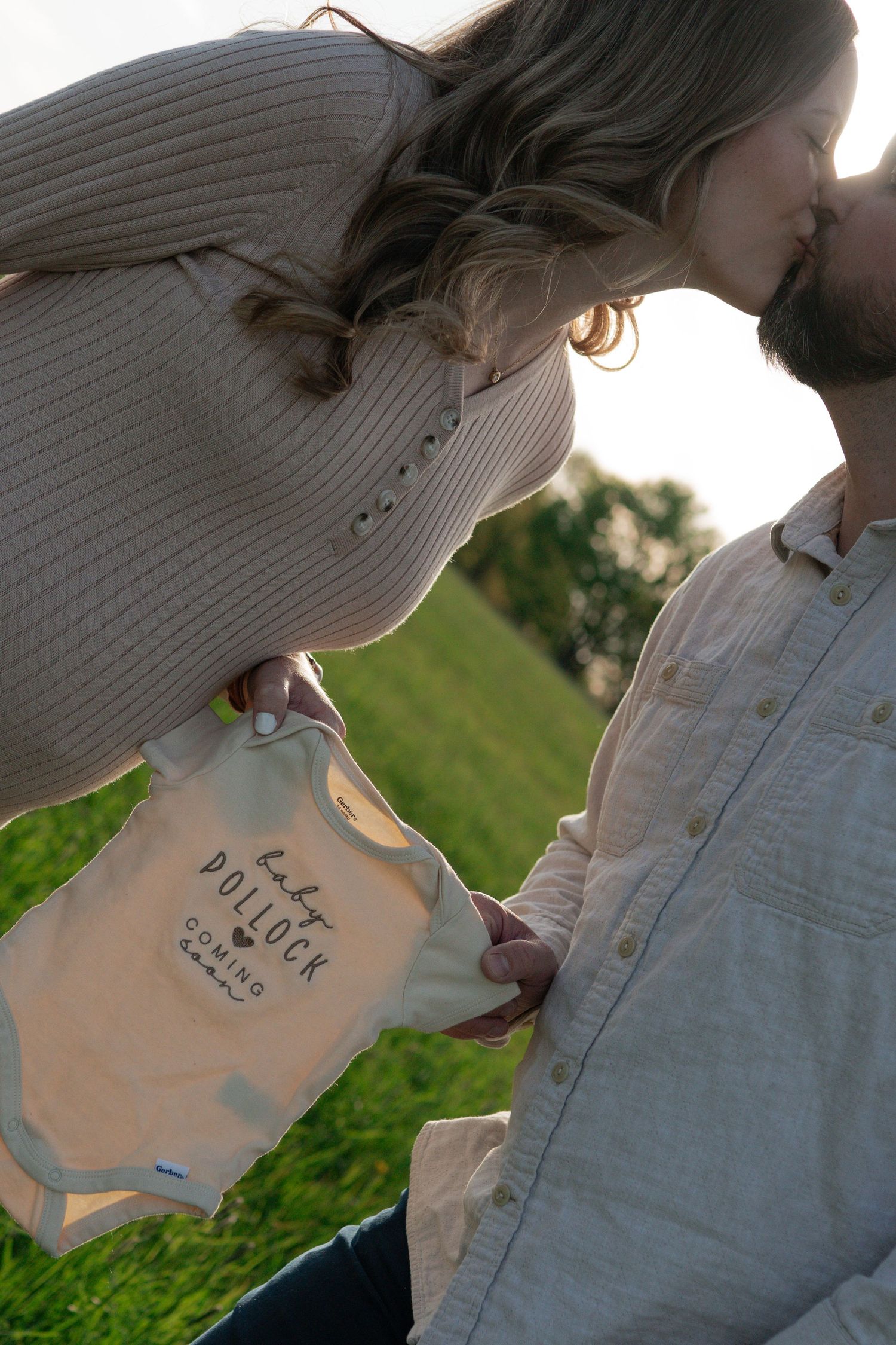 Couple kissing outdoors holding a white baby onesie reading Baby Pollock Coming Soon for pregnancy announcement.