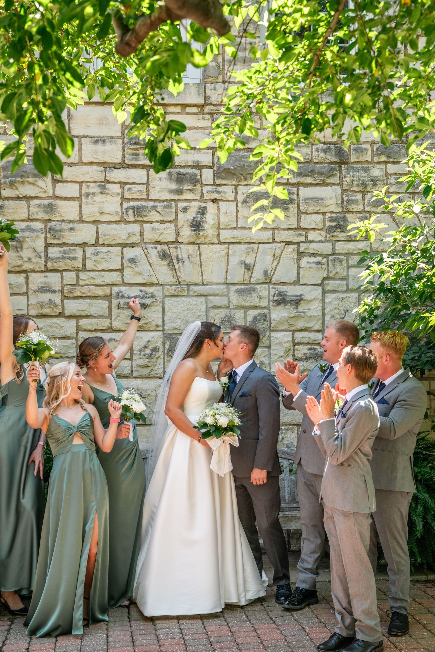Wedding party celebrates with sparklers against stone wall backdrop under green foliage on sunny day.