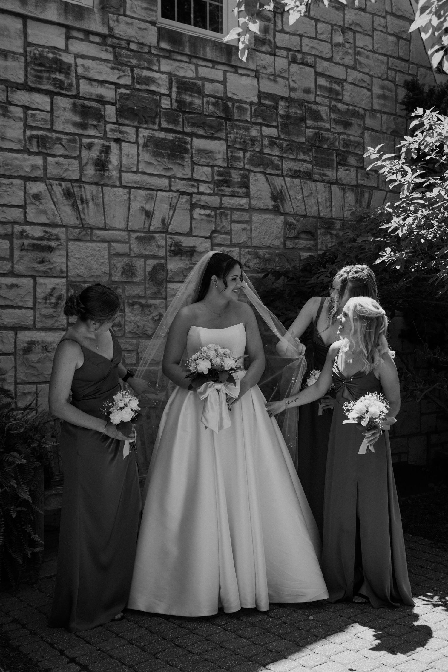 Bride in white gown with bridesmaids in gray dresses posing by stone wall.