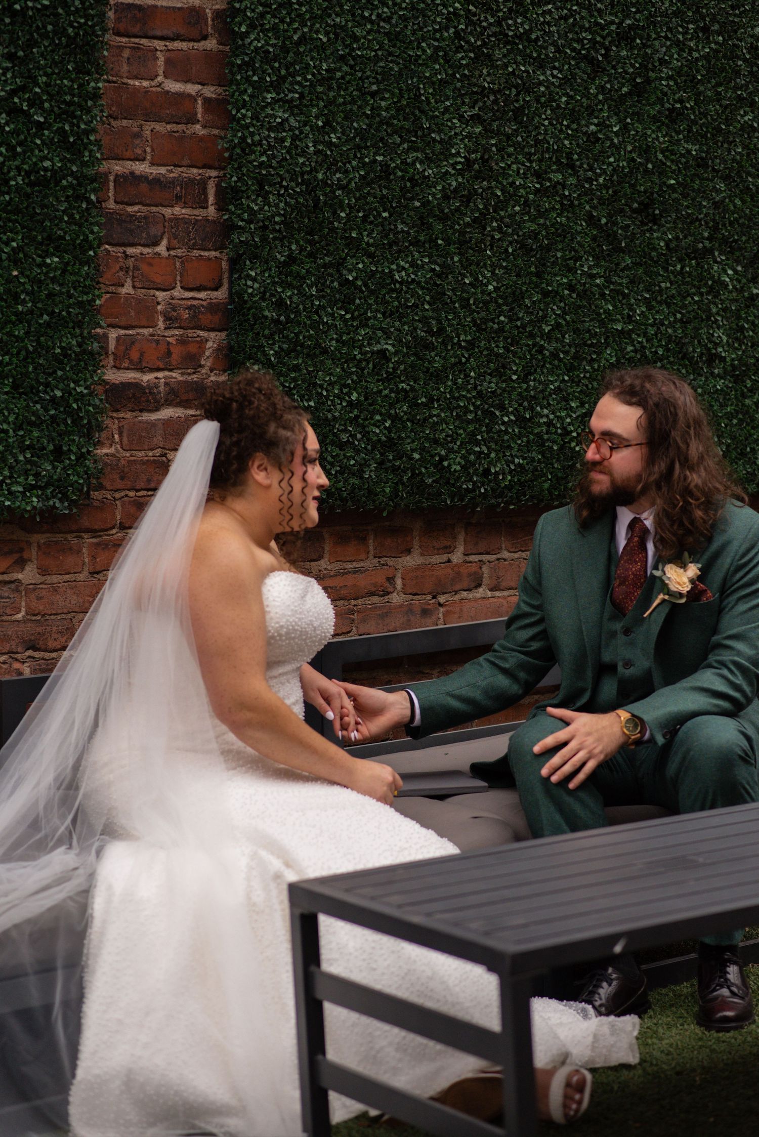 Bride in white gown and veil holds hands with groom in green suit, sitting together against brick and hedge wall.