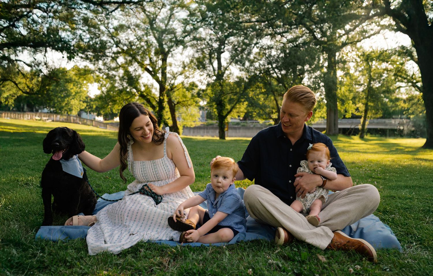 Happy family with young children and a black dog enjoying a sunny day on a blanket in a lush green park.