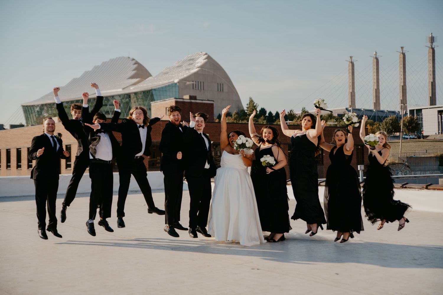 Wedding party in black attire jumping joyfully on rooftop with bride in white gown holding bouquet on sunny day.