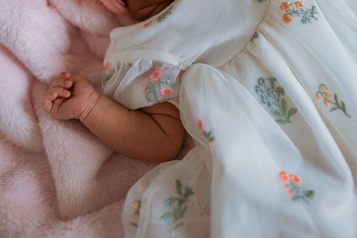Sleeping newborn baby in a delicate white floral embroidered dress on a soft pink blanket.