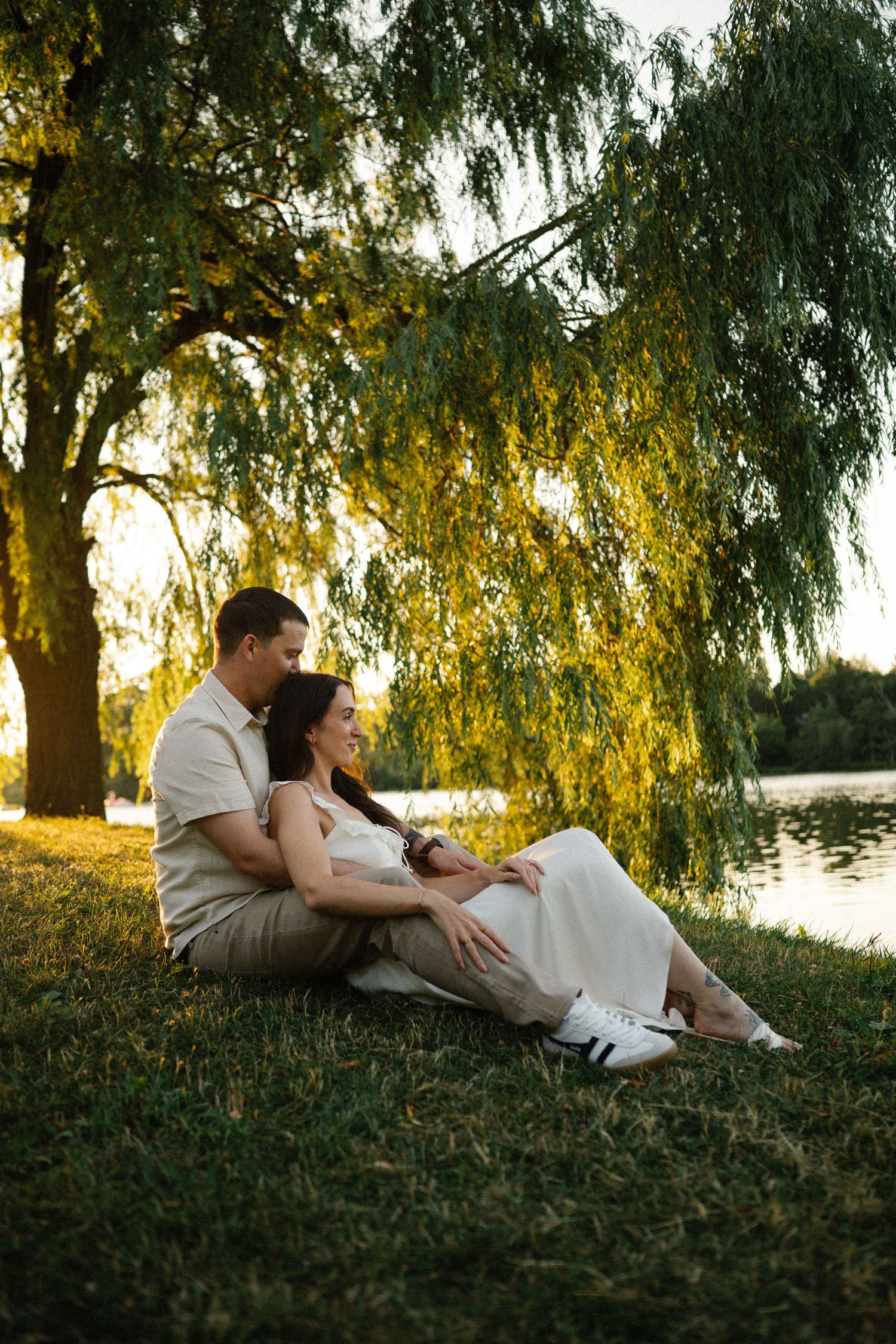 Couple sitting together on grass under a weeping willow tree by a lake during golden hour sunset.