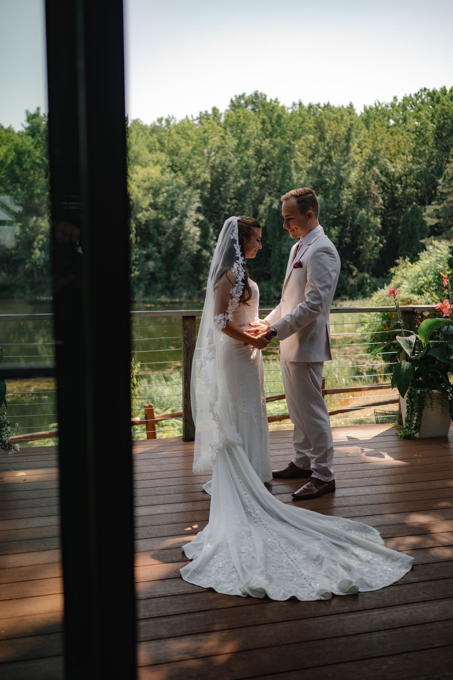 Bride and groom hold hands on a wooden deck surrounded by lush greenery during an outdoor wedding ceremony.