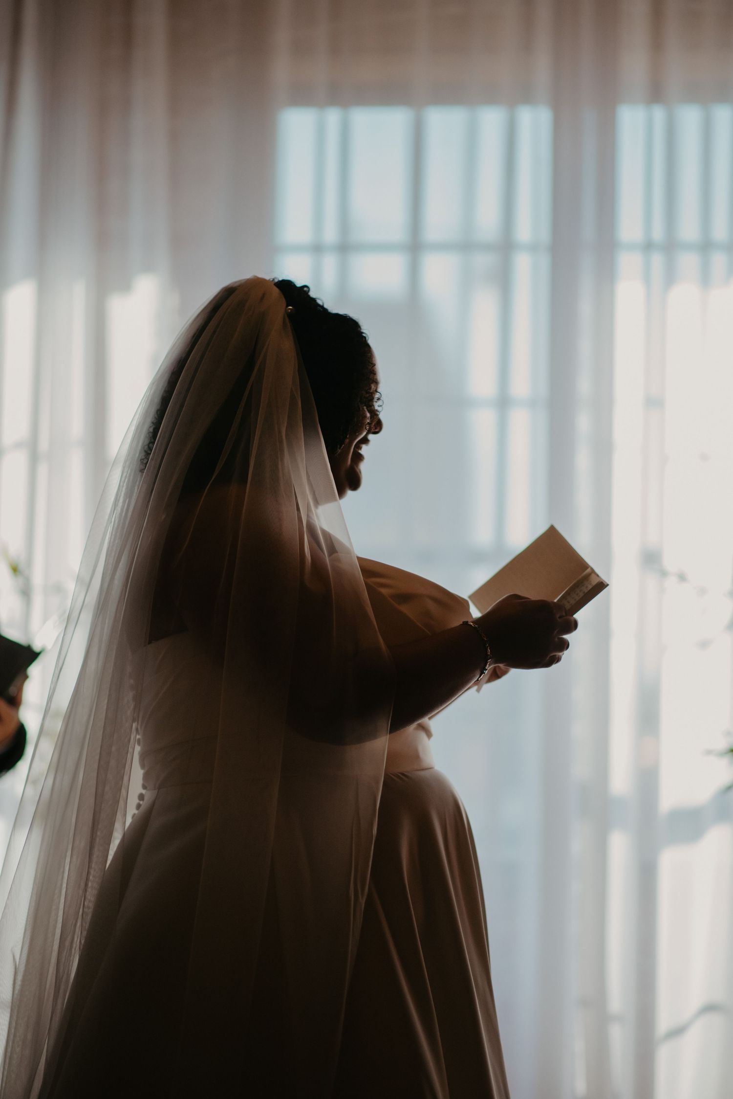 Bride in white gown and veil reads a letter by a sunlit window in a romantic silhouette wedding photo.