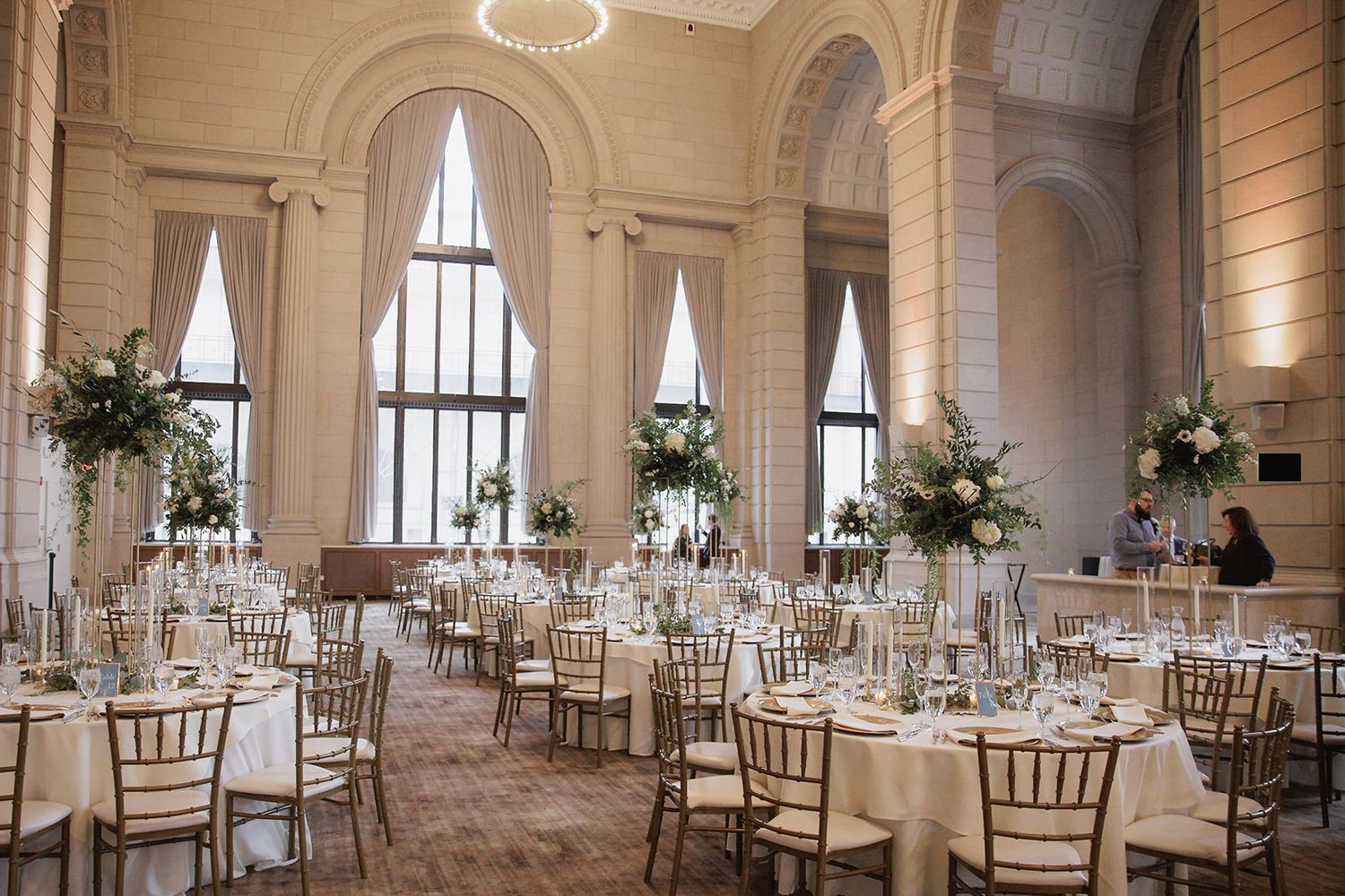 Elegant ballroom wedding reception with tall floral centerpieces, white linens and gold chiavari chairs in historic venue.