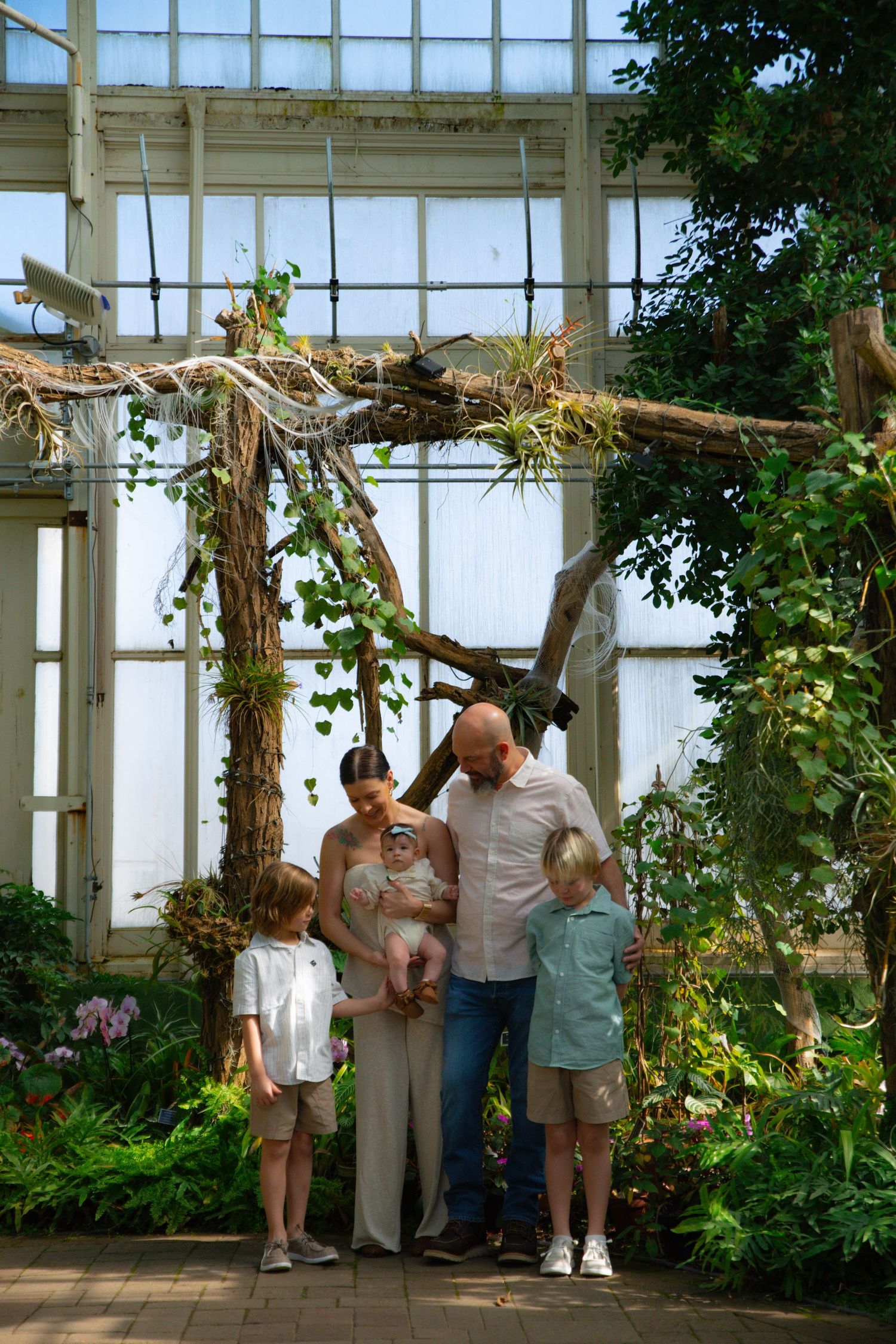 Family of five posing together in a lush greenhouse conservatory beneath a rustic driftwood arch.