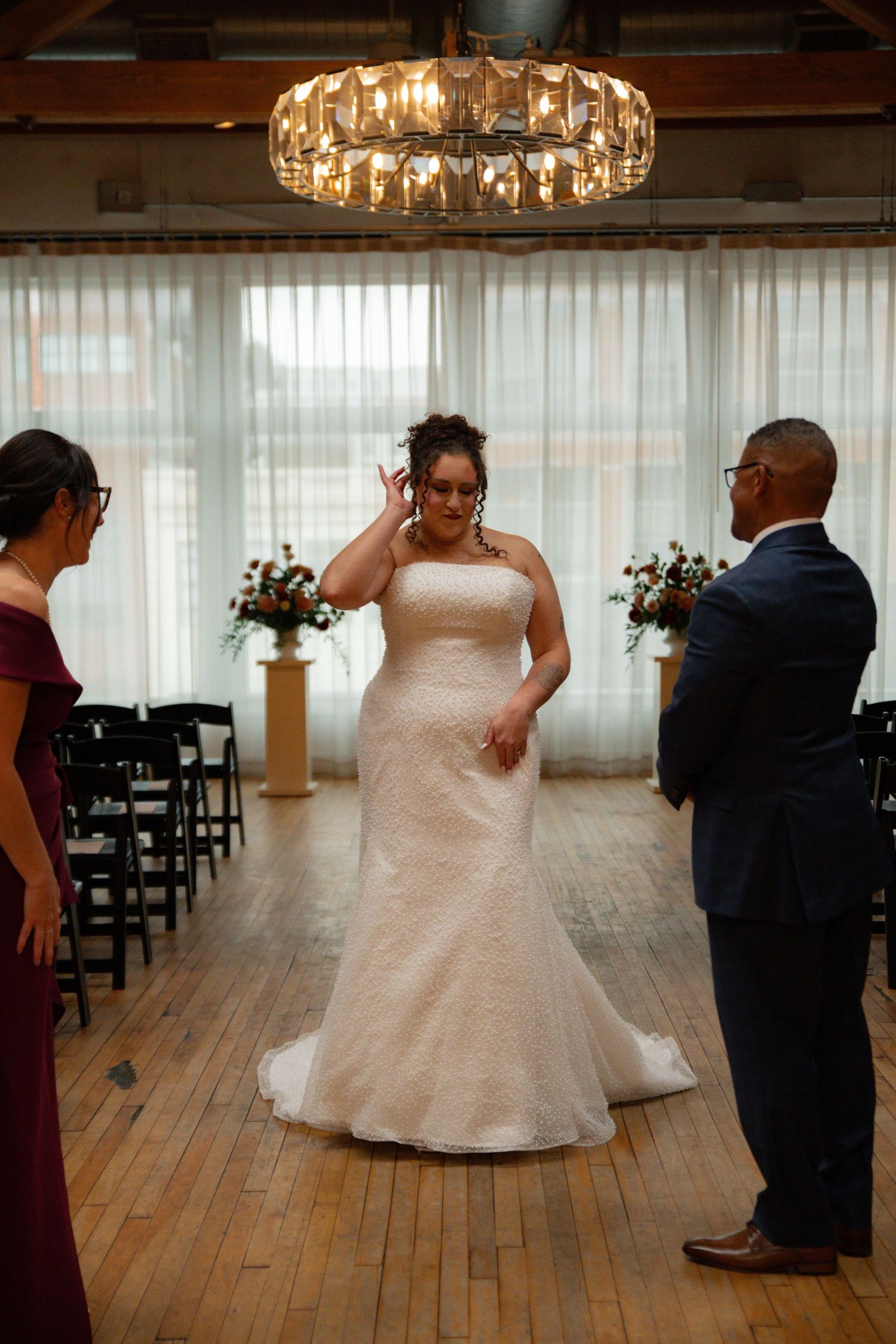 Bride in strapless white gown walks down aisle at indoor wedding ceremony with chandelier overhead.