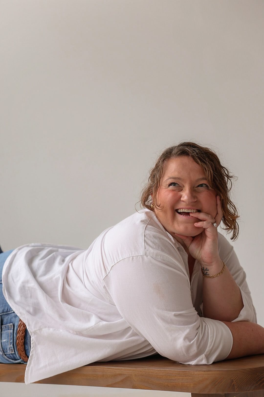 A cheerful plus-size woman in a white shirt and jeans lies on a wooden bench, smiling warmly in a bright studio.